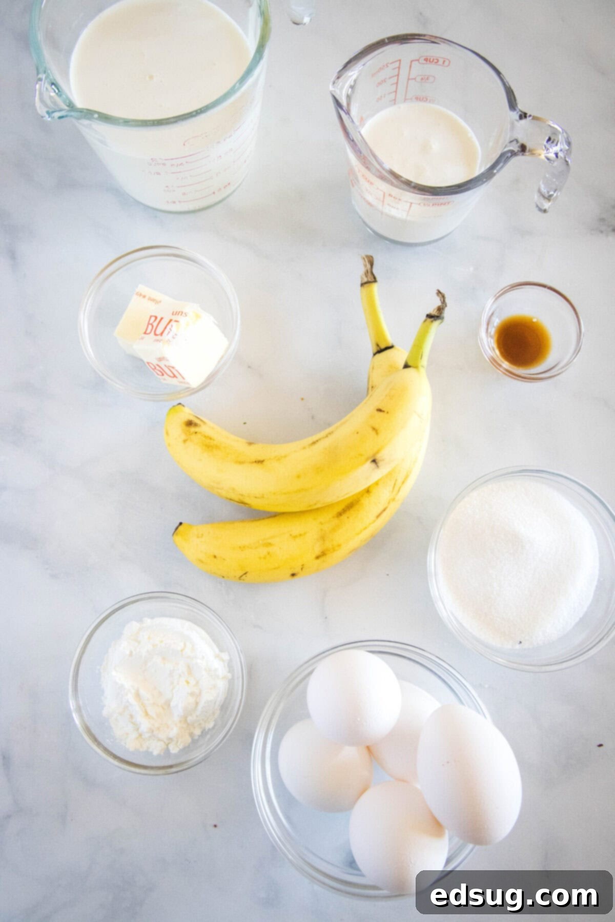 Overhead view of all the key ingredients for making banana cream pie neatly arranged: bowls of egg yolks, granulated sugar, cornstarch, unsalted butter, whole milk, heavy cream, vanilla extract, and two perfectly ripe bananas.