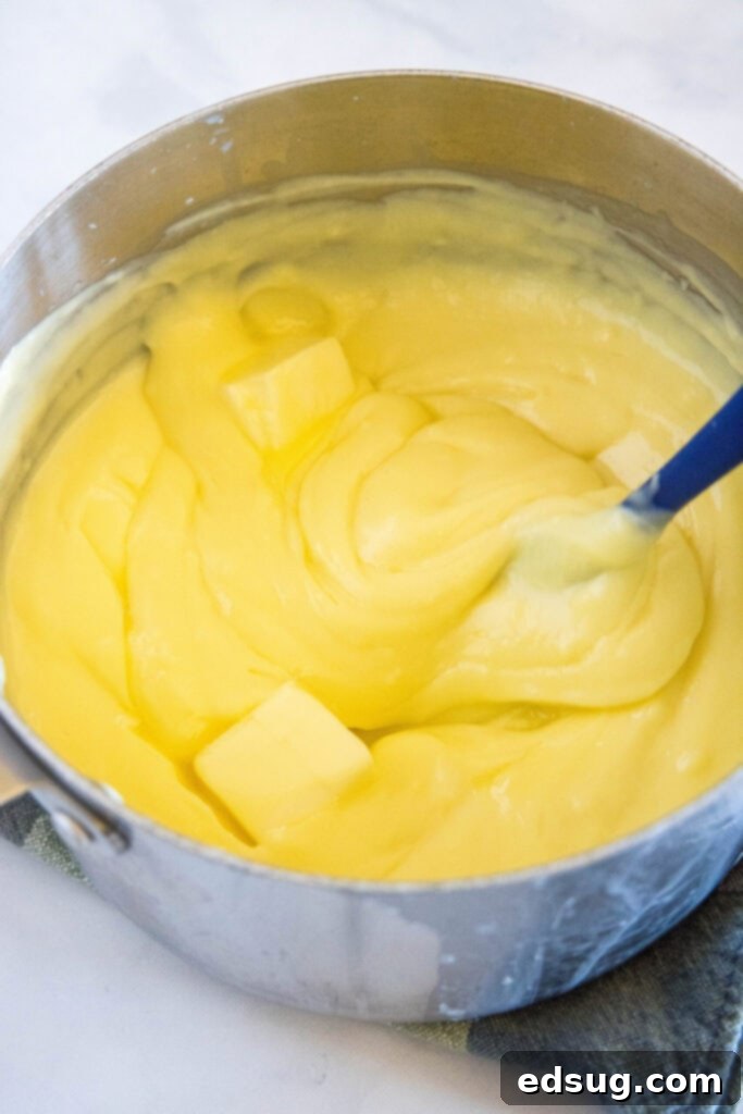 A spatula gently stirring cubes of butter into the warm, thick banana cream pie custard in a saucepan, creating a smooth and glossy finish.