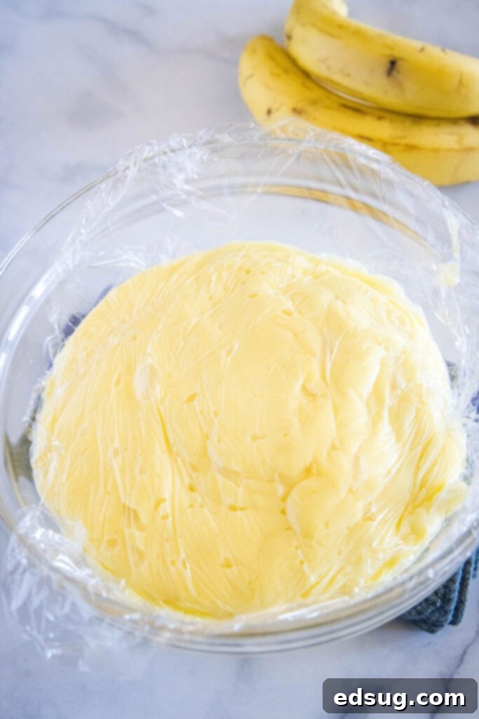 A bowl of rich, smooth banana cream pie filling, perfectly covered with plastic wrap pressed directly onto its surface to prevent skin formation, chilling in the refrigerator.