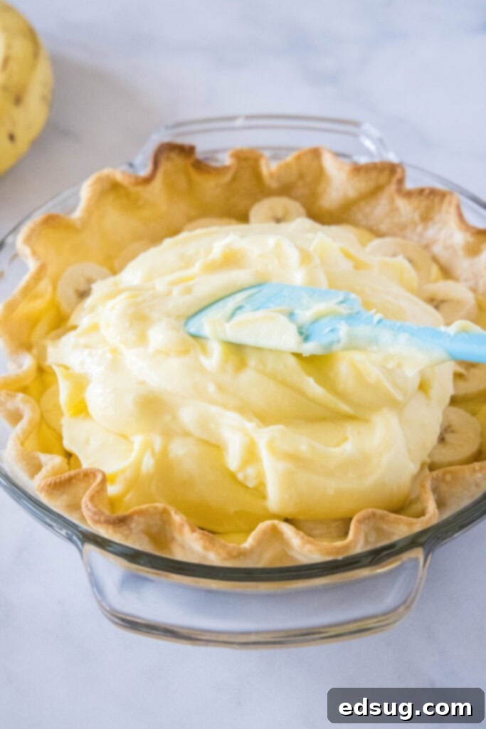 A spatula smoothly spreading the thick, creamy banana custard filling into a pre-baked pastry shell, ensuring an even layer over the bananas.