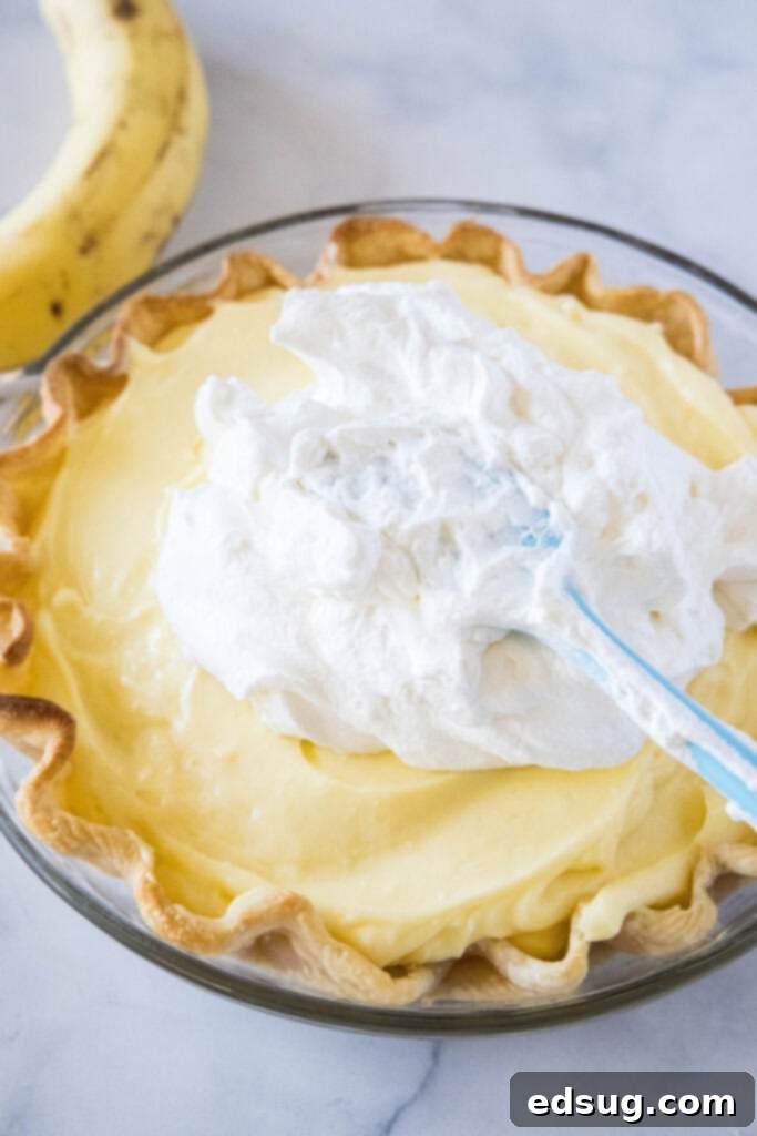 A spatula gracefully spreading fluffy, white whipped cream over the golden-brown surface of a finished banana cream pie, creating a smooth and inviting topping.