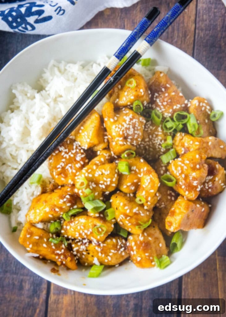 An overhead shot of a white bowl filled with Bourbon Chicken, rice, green onions, and sesame seeds