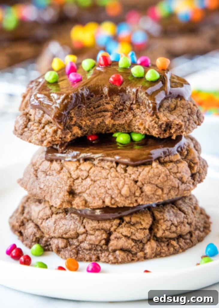 Stellar Brownie Cookies 2 Stack of four thick, fudgy cosmic brownie cookies with a bite missing from the top one, revealing a soft interior and topped with chocolate ganache and colorful rainbow sprinkles.