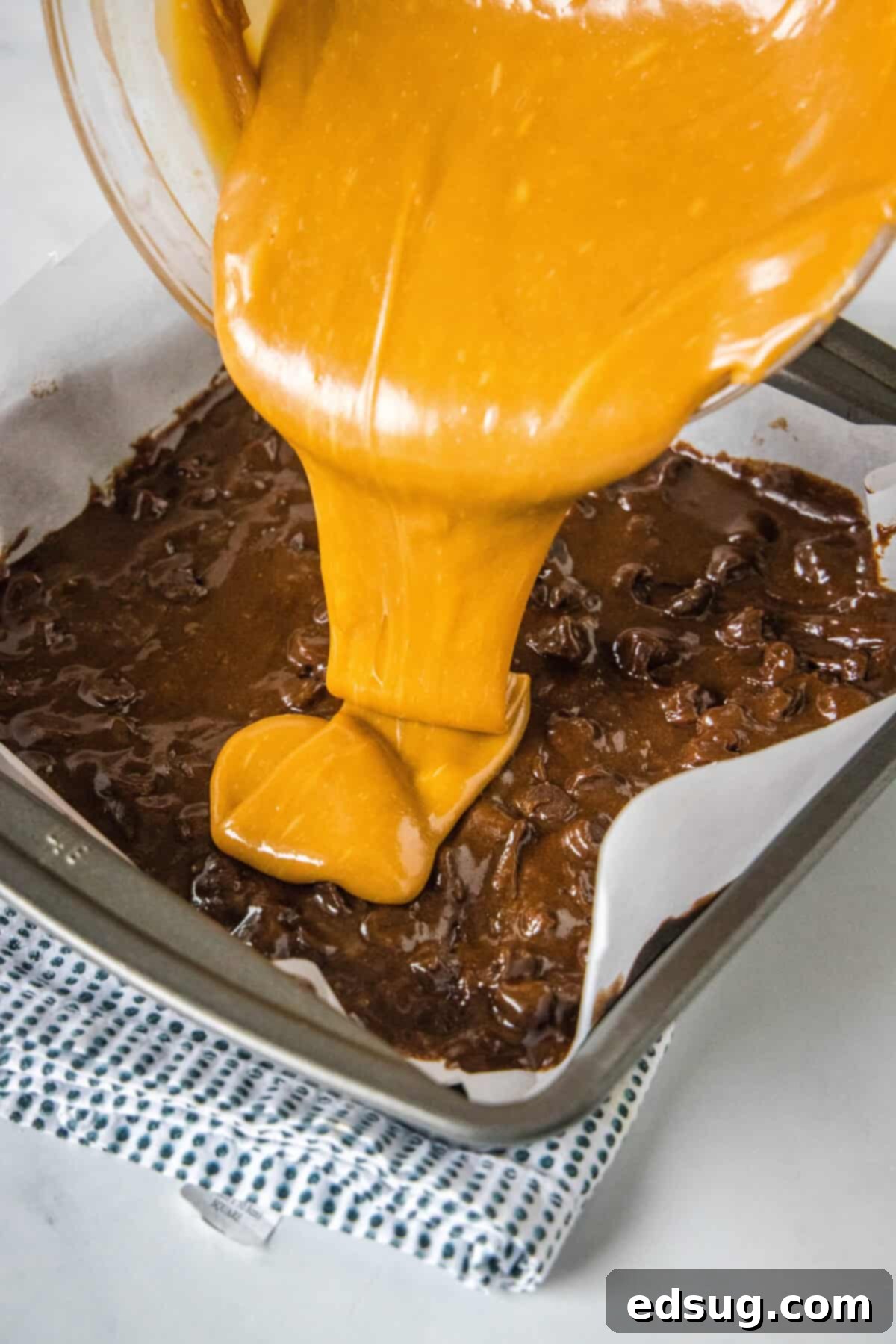 Watch the magic happen as gooey salted caramel blankets the rich chocolate brownie base. Smooth, melted salted caramel being generously poured over a layer of chocolate brownie batter in a parchment-lined square baking pan.