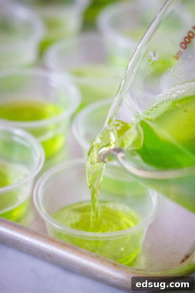 Witches Brew Jello Shots 3 Carefully pouring a vibrant green Jell-O mixture into small disposable cups on a baking tray to form the first layer of Halloween Jello Shots.