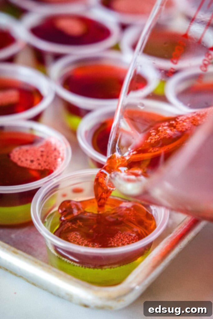 Merry & Bright Jello Shots 5 Carefully pouring a layer of red liquid JELL-O over a set green JELL-O layer in small plastic cups for a festive, two-toned Christmas shot.