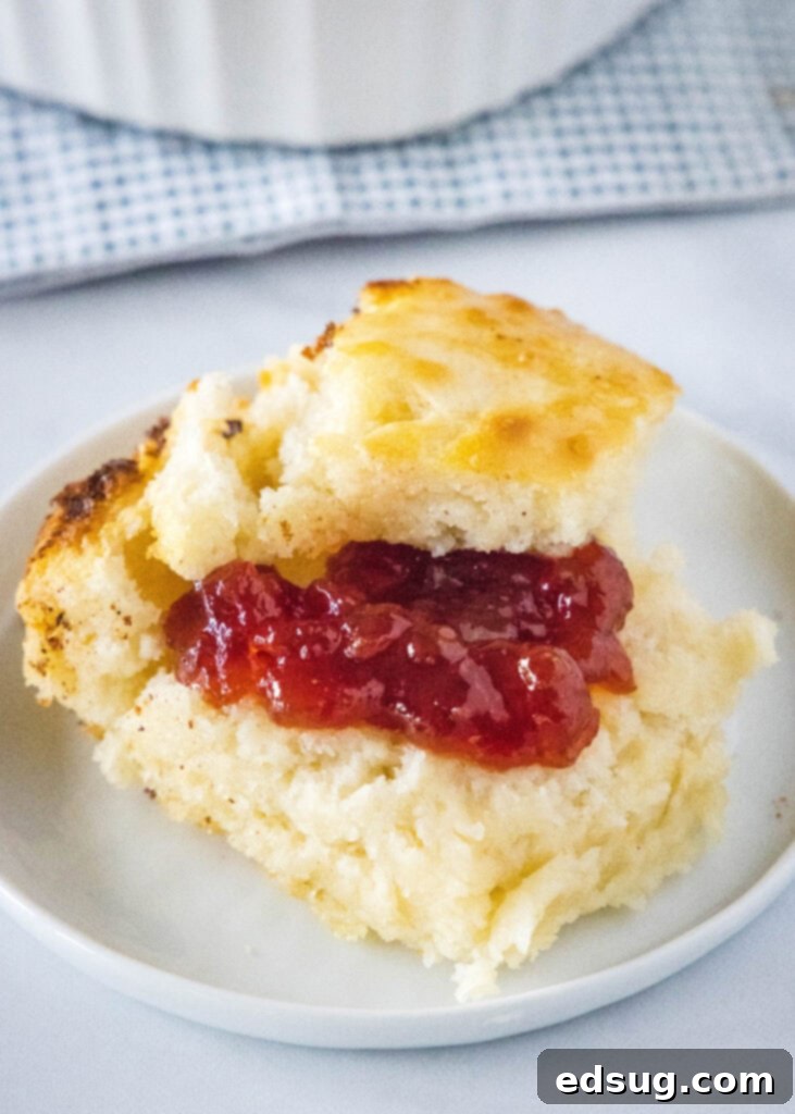 A golden-brown butter swim biscuit topped with a dollop of vibrant red jelly, resting on a pristine white plate, showcasing its soft texture and buttery aroma.