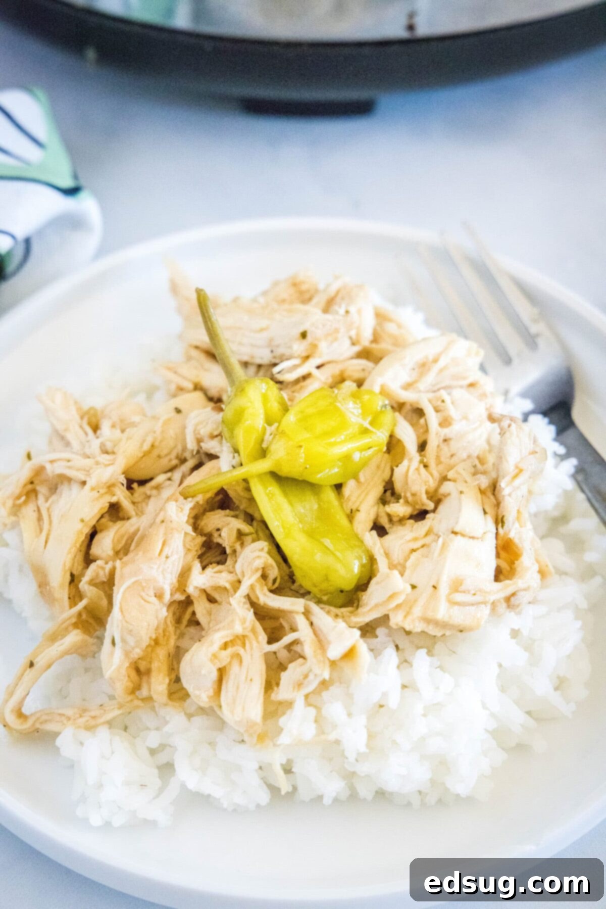 This slow cooker Mississippi chicken recipe is juicy, tender, and full of rich ranch and onion soup mix, with a kick of spice. A plate with a fork and a bed of rice, topped with shredded Mississippi chicken and vibrant pepperoncini peppers