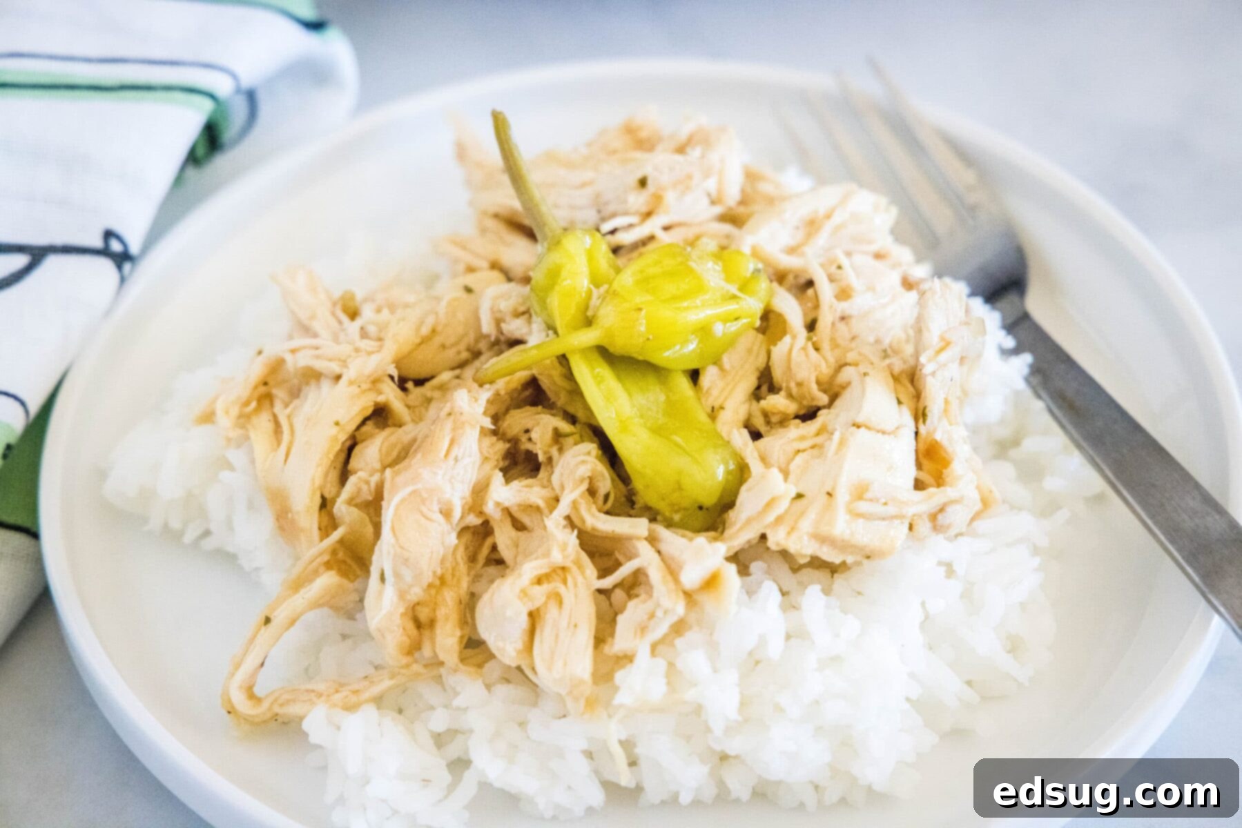 This slow cooker Mississippi chicken recipe is juicy, tender, and full of rich ranch and onion soup mix, with a kick of spice. A plate with a fork and some rice, topped with shredded chicken and pepperoncini peppers, next to a kitchen towel