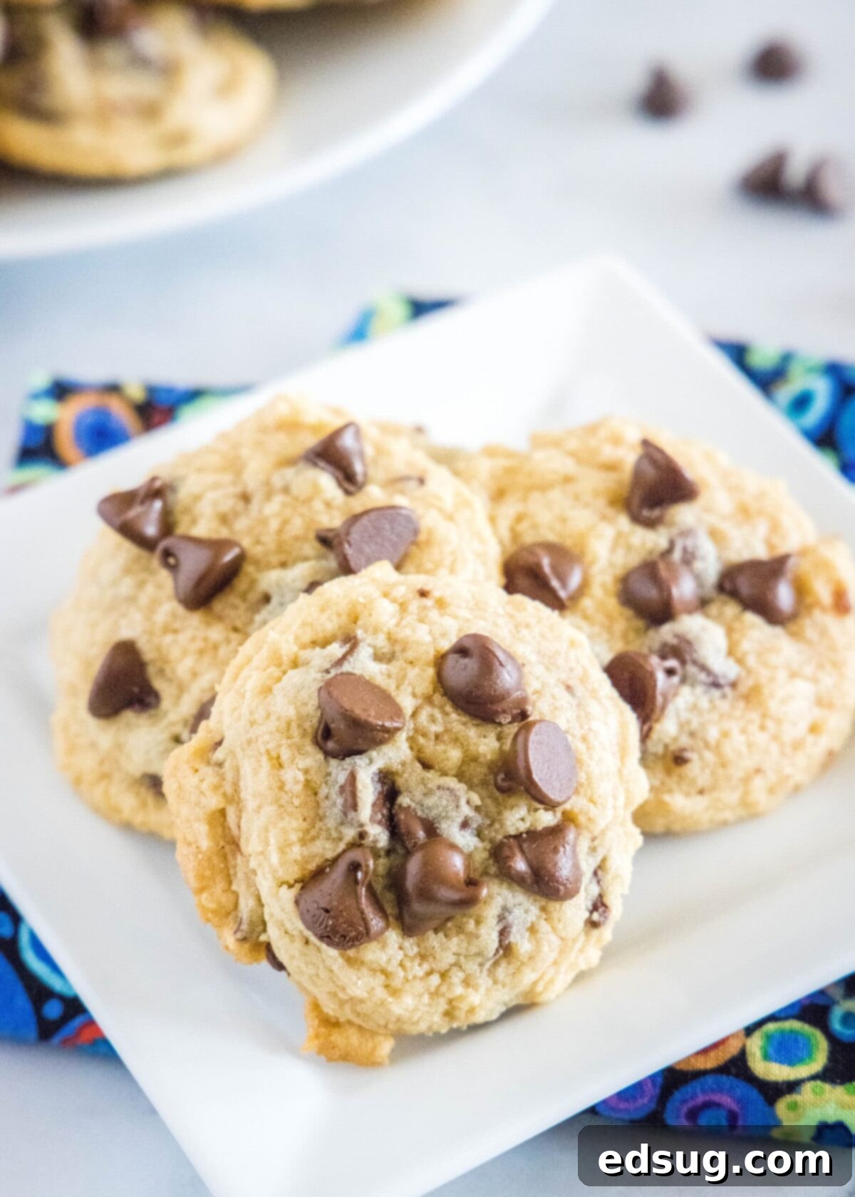 Close up of three chocolate chip cookies on a white plate on top of a kitchen towel, with cookies and chocolate chips in the background