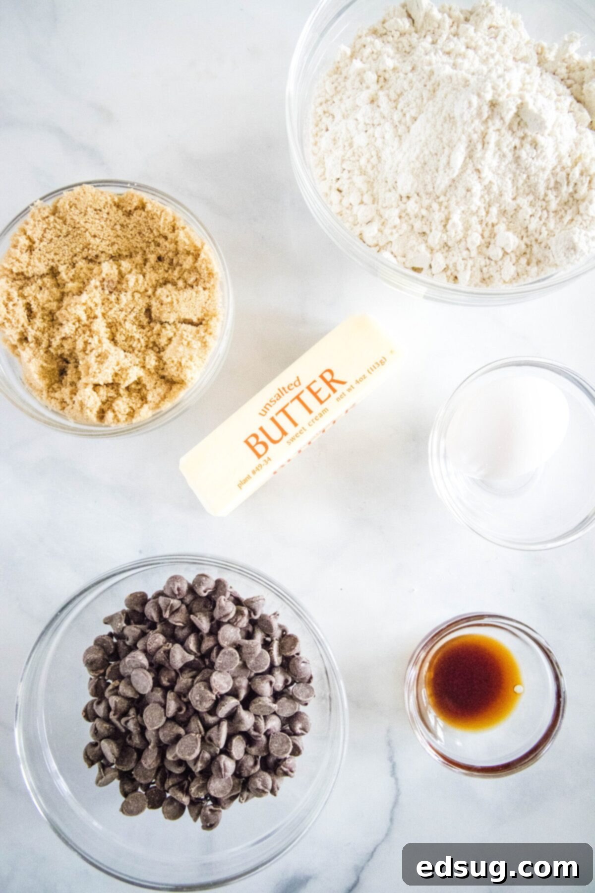 Overhead view of the ingredients needed for Bisquick cookies: A bowl of Bisquick, a bowl of brown sugar, a bowl of chocolate chips, a stick of butter, a ramekin of vanilla, and a bowl with an egg
