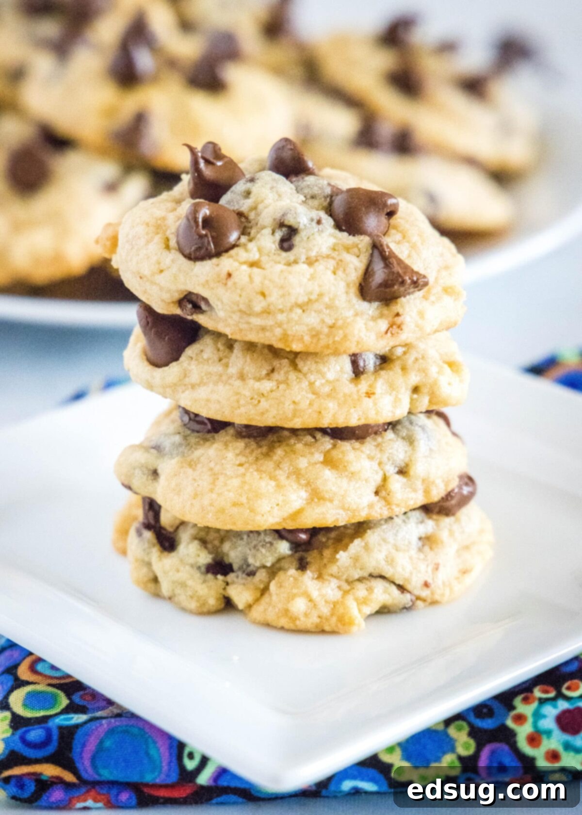 Four chocolate chip cookies stacked on a plate, with more cookies in the background