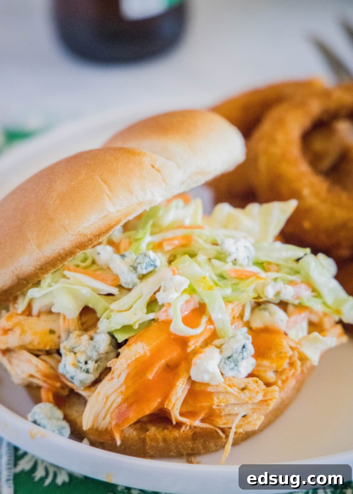 A chicken sandwich topped with coleslaw and blue cheese, on a plate, with onion rings in the background