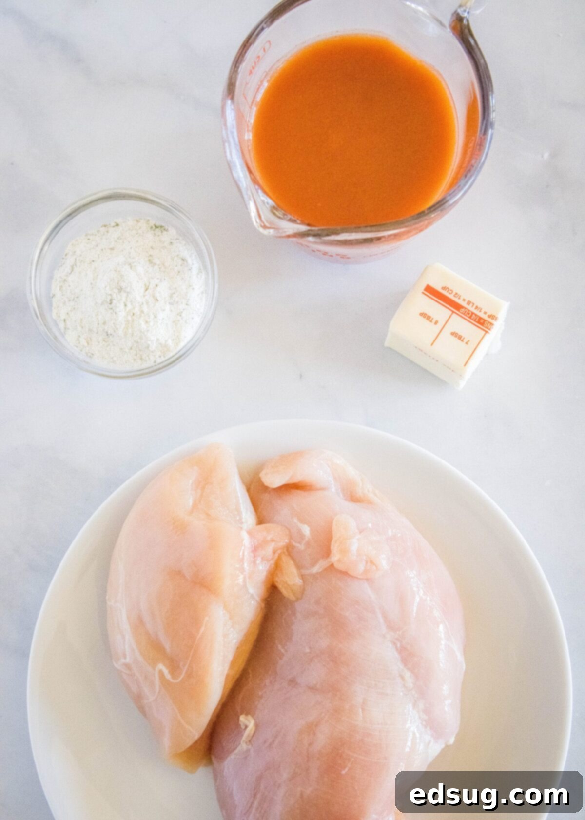 Overhead view of a knob of butter, a plate of raw chicken breasts, a ramekin of ranch dressing mix, and a pyrex of buffalo sauce