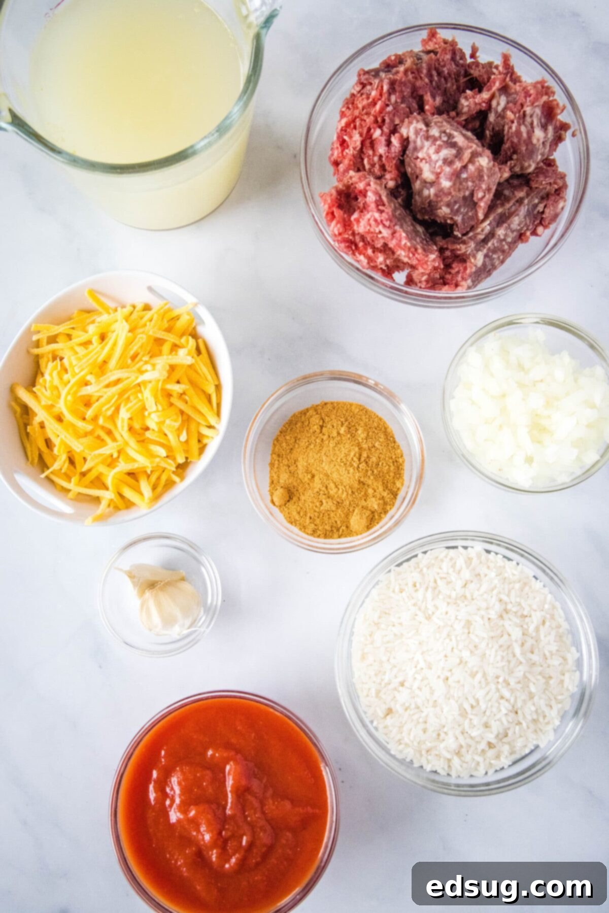 Overhead view of the ingredients needed for taco rice: a bowl of raw ground beef, a bowl of shredded cheddar cheese, a bowl of rice, a bowl of taco seasoning, a bowl of diced onions, a bowl of garlic, a bowl of tomato sauce, and a pitcher of chicken broth