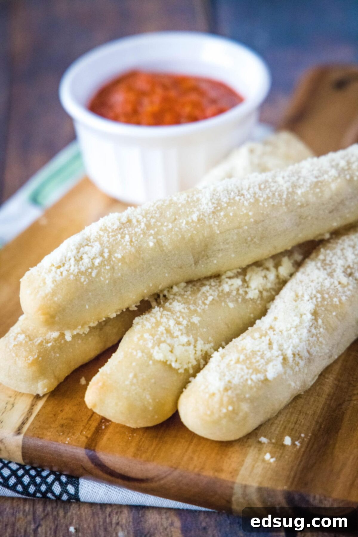 Four golden-brown homemade crazy breadsticks on a wooden cutting board, with a small ramekin of marinara sauce in the soft-focus background, ready for dipping.