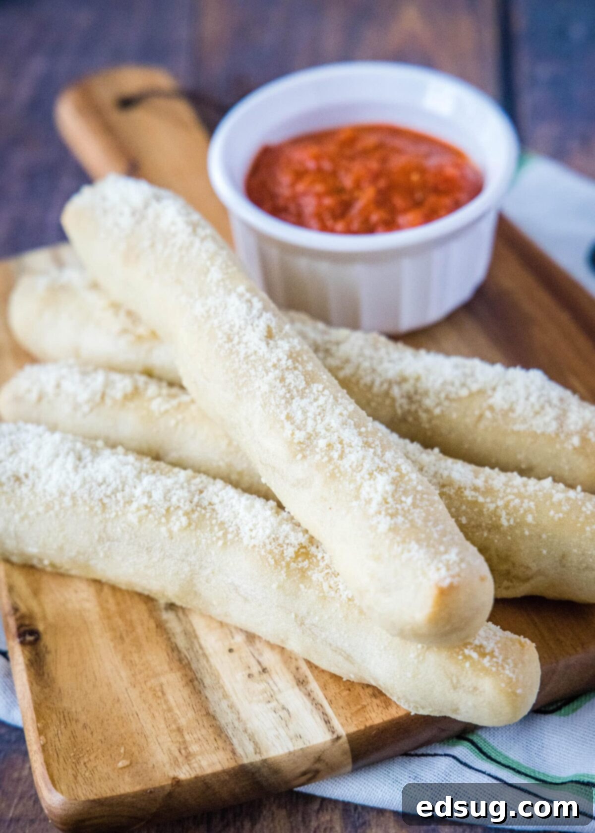 A stack of four perfectly baked and cheesy breadsticks on a wooden cutting board, with a small white ramekin of marinara sauce in front, all against a clean backdrop.