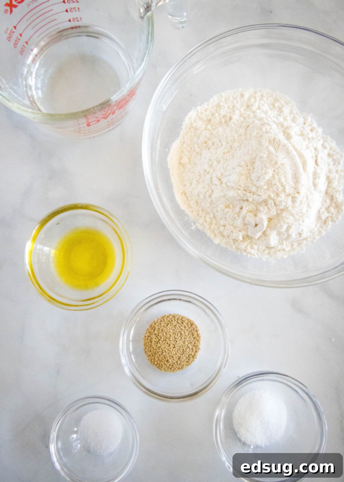 Overhead shot of all the dry and wet ingredients laid out on a table for making pizza dough: a bowl of all-purpose flour, a small bowl of active dry yeast, a small bowl of sugar, a small bowl of salt, a bottle of olive oil, and a glass of warm water in a Pyrex measuring cup.