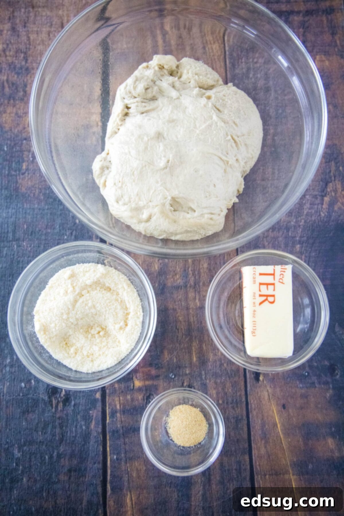 Close-up overhead view of the ingredients for the crazy bread topping: a bowl of freshly made pizza dough, a bowl of grated Parmesan cheese, a bowl of garlic powder, and half a stick of softened butter.