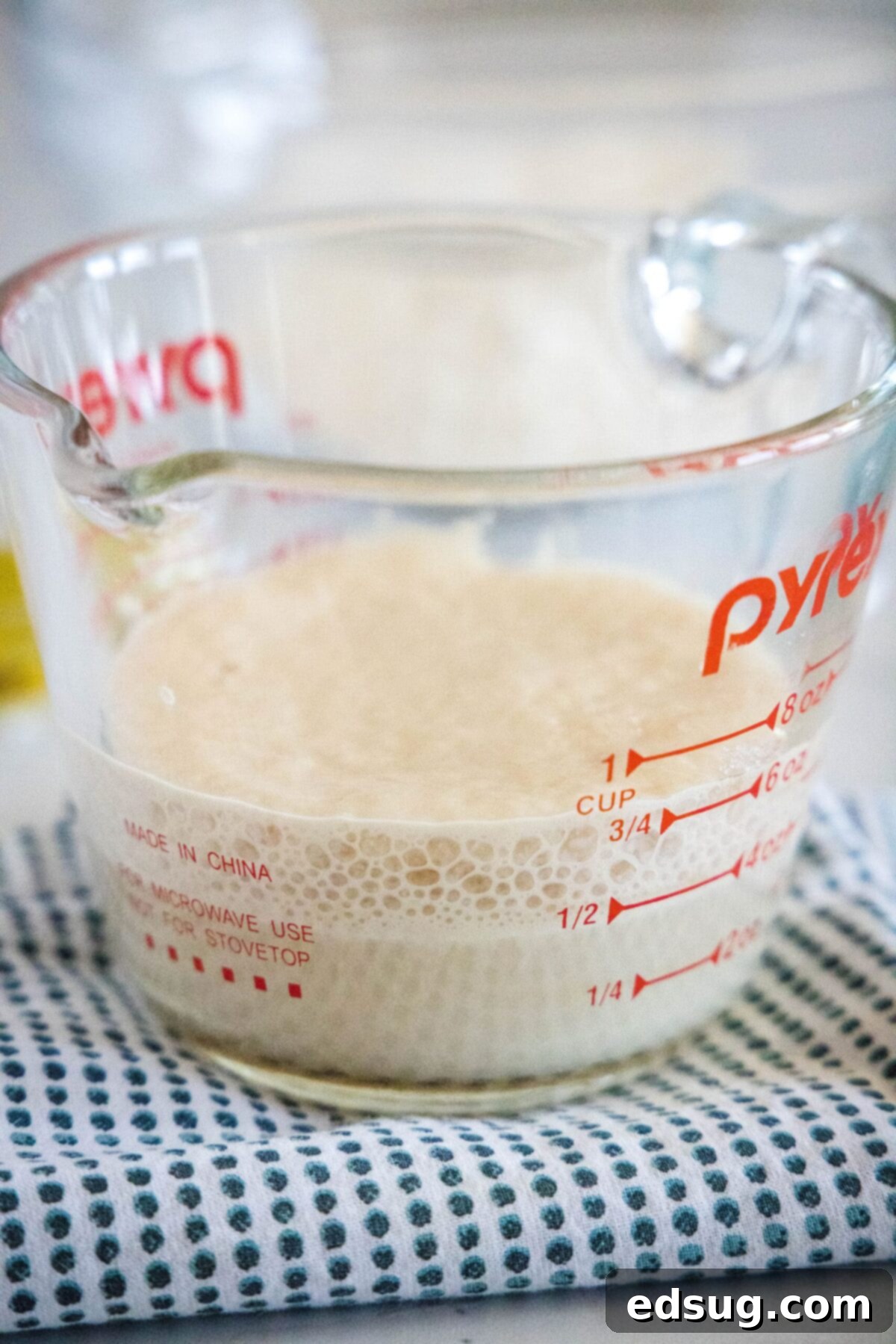 A clear Pyrex measuring cup with warm water, sugar, and active dry yeast, showing a foamy, bubbly layer on top, indicating the yeast has bloomed successfully.