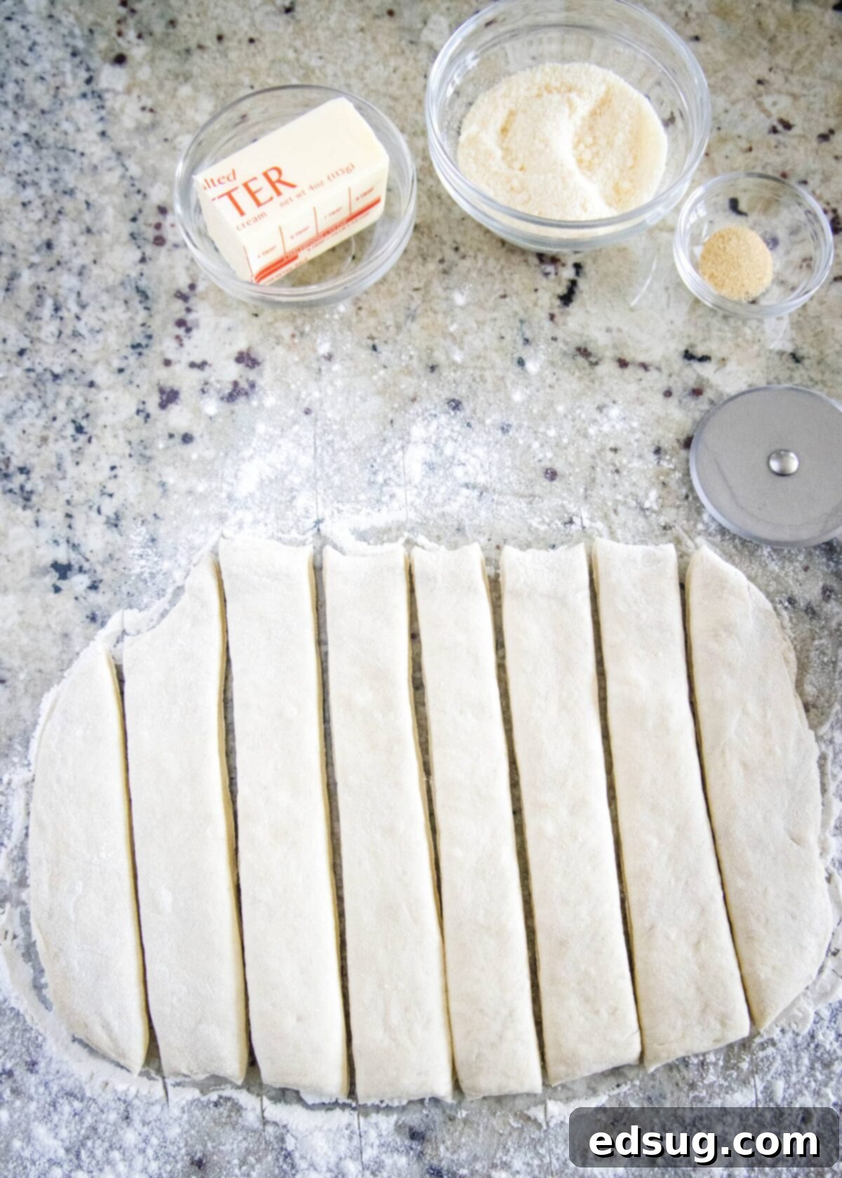 Eight strips of pizza dough arranged neatly on a wooden cutting board, alongside a pizza cutter, a bowl of grated Parmesan cheese, a bowl of garlic powder, and half a stick of butter, ready for shaping and baking.