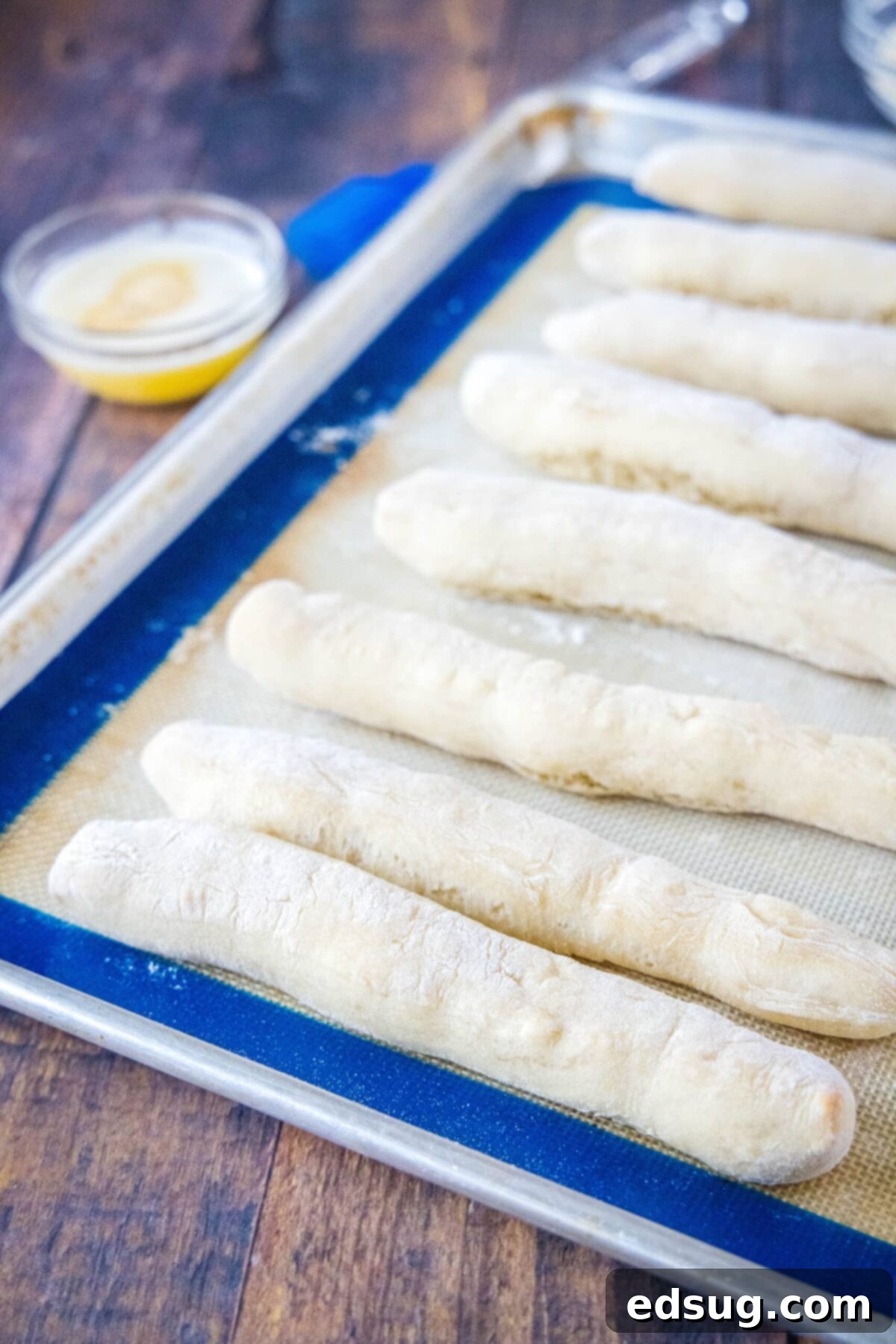 Freshly baked golden-brown breadsticks cooling slightly on a baking sheet lined with parchment paper, with a bowl of melted butter in the background.