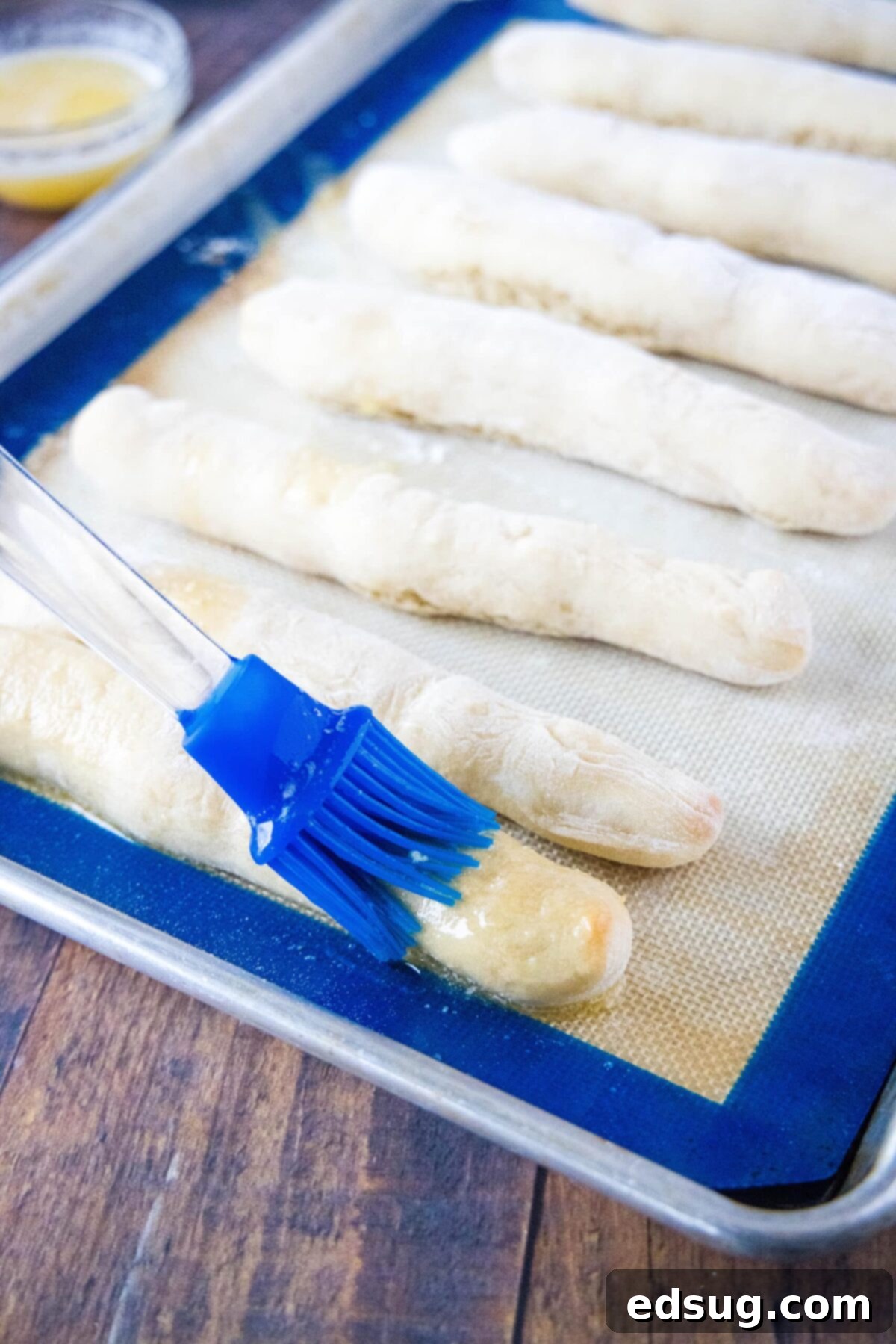 A kitchen brush gracefully applying melted garlic butter onto warm, golden-brown breadsticks directly on the baking sheet, ensuring every piece is coated.