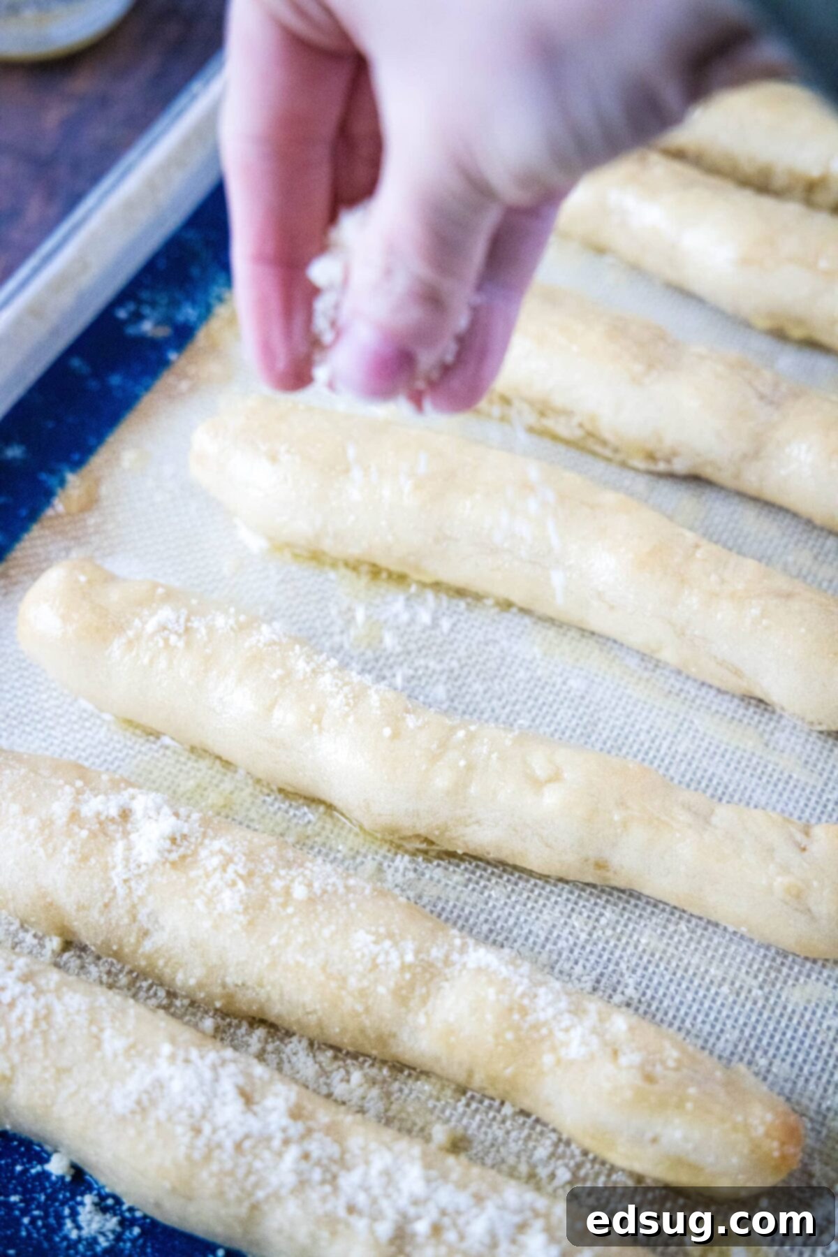 A hand carefully sprinkling grated Parmesan cheese over the warm, buttered breadsticks arranged on a baking sheet, creating the final cheesy layer.