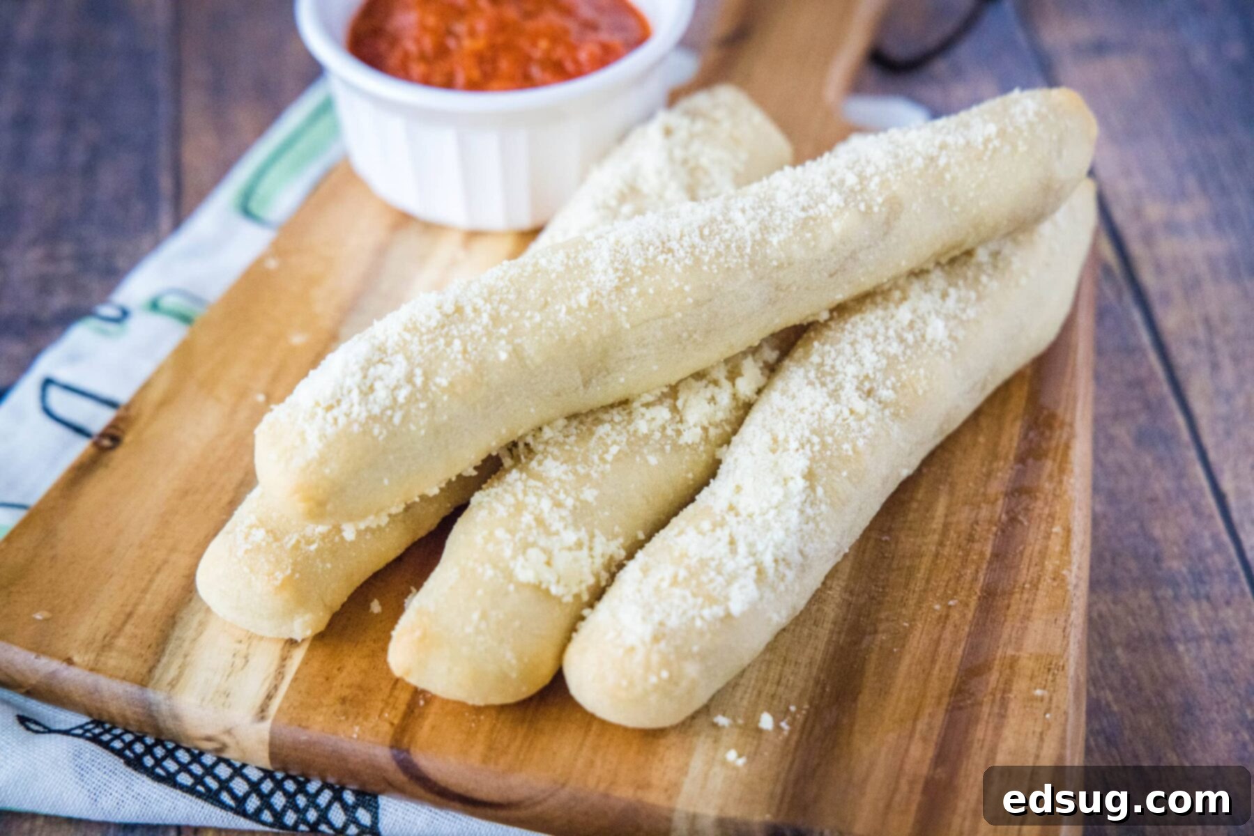 A tempting pile of four golden-brown crazy breadsticks resting on a rustic wooden cutting board, with a small ramekin of vibrant marinara sauce poised for dipping in the background.