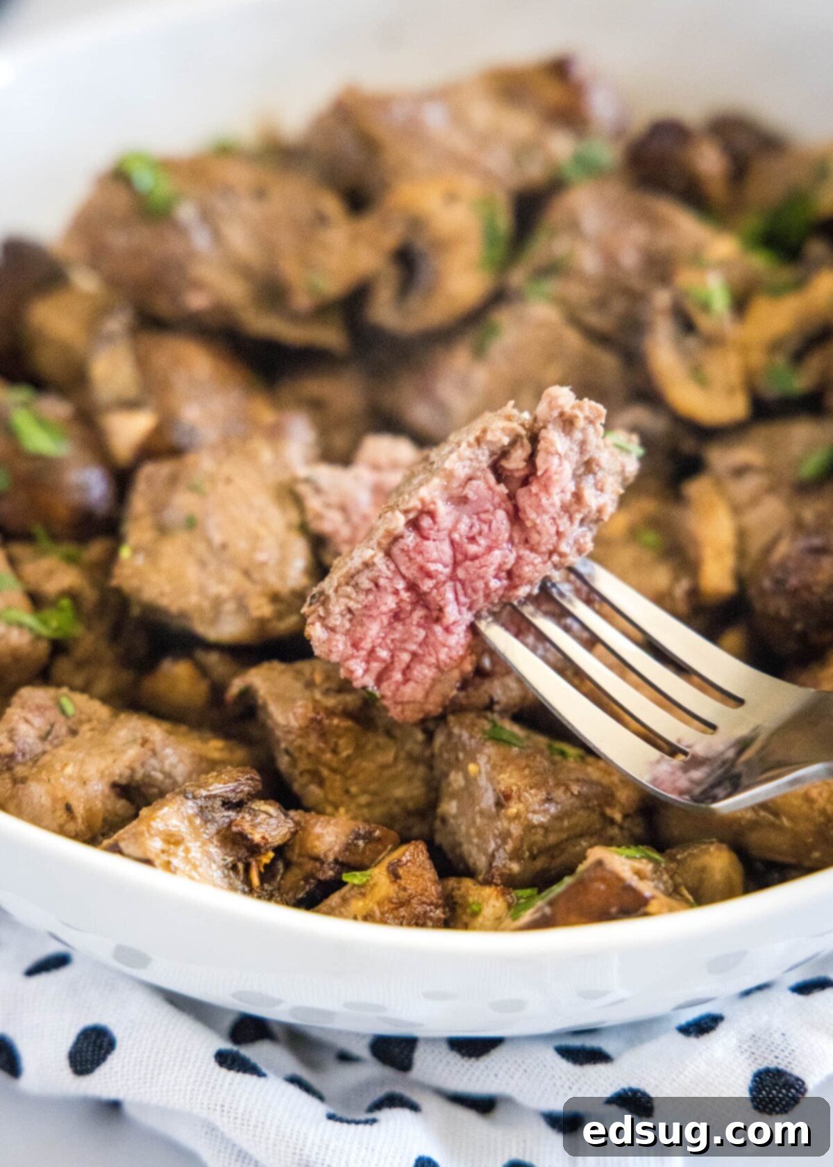 How to make easy air fryer steak bites with mushrooms and garlic powder, resulting in tender meat and juicy mushrooms in just 15 minutes! A succulent piece of steak on a fork, with a plate of golden-brown steak and mushroom bites served in the background, showcasing their tender texture and rich color.