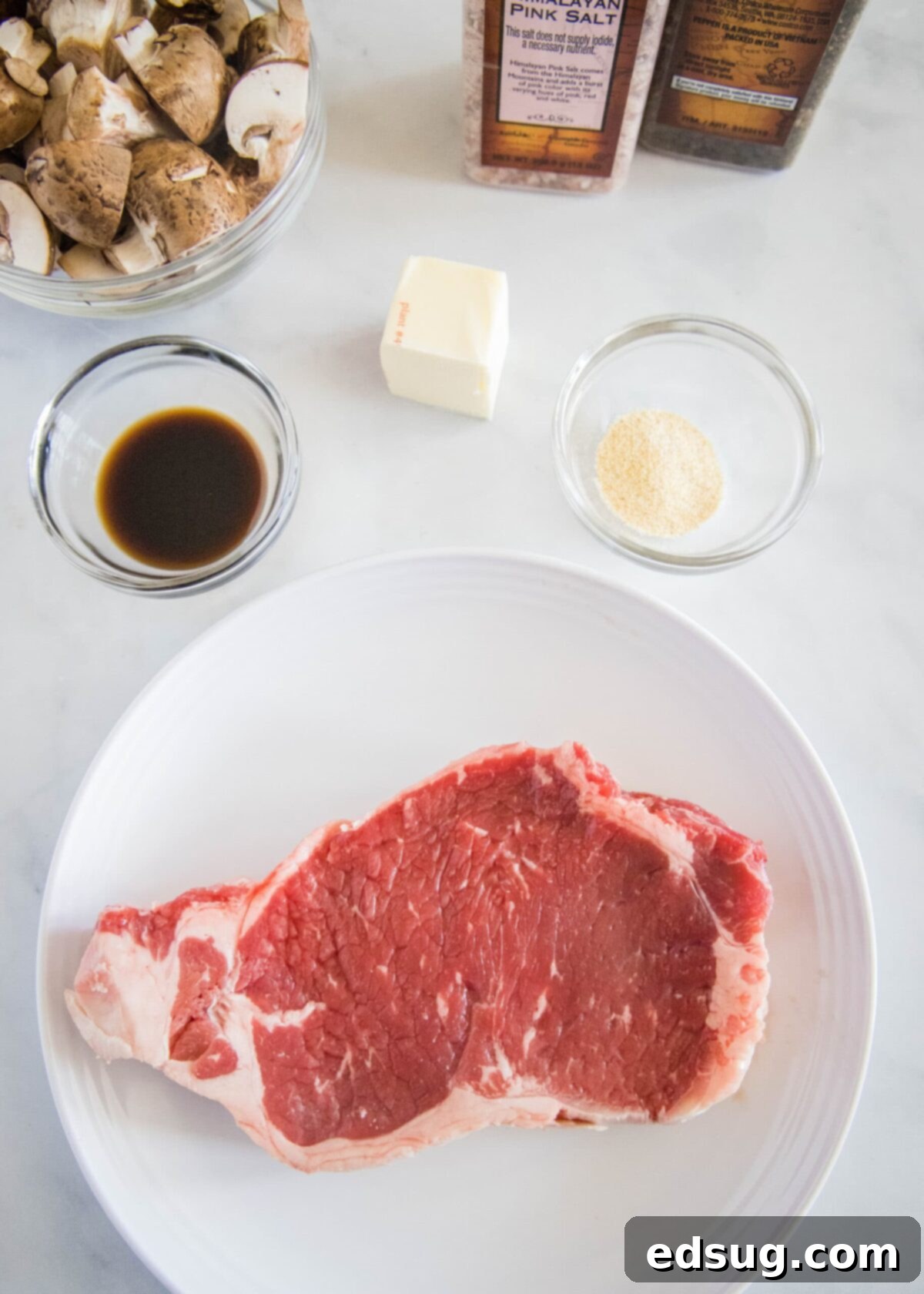 How to make easy air fryer steak bites with mushrooms and garlic powder, with tender meat and juicy mushrooms. It only takes 15 minutes! An overhead shot showcasing all the fresh and essential ingredients required for making delicious air fryer steak and mushroom bites: a raw sirloin steak on a white plate, a bowl brimming with fresh brown mushrooms, a pat of rich butter, a small dish of aromatic garlic powder, a bottle of savory Worcestershire sauce, and shakers of coarse salt and freshly ground black pepper.