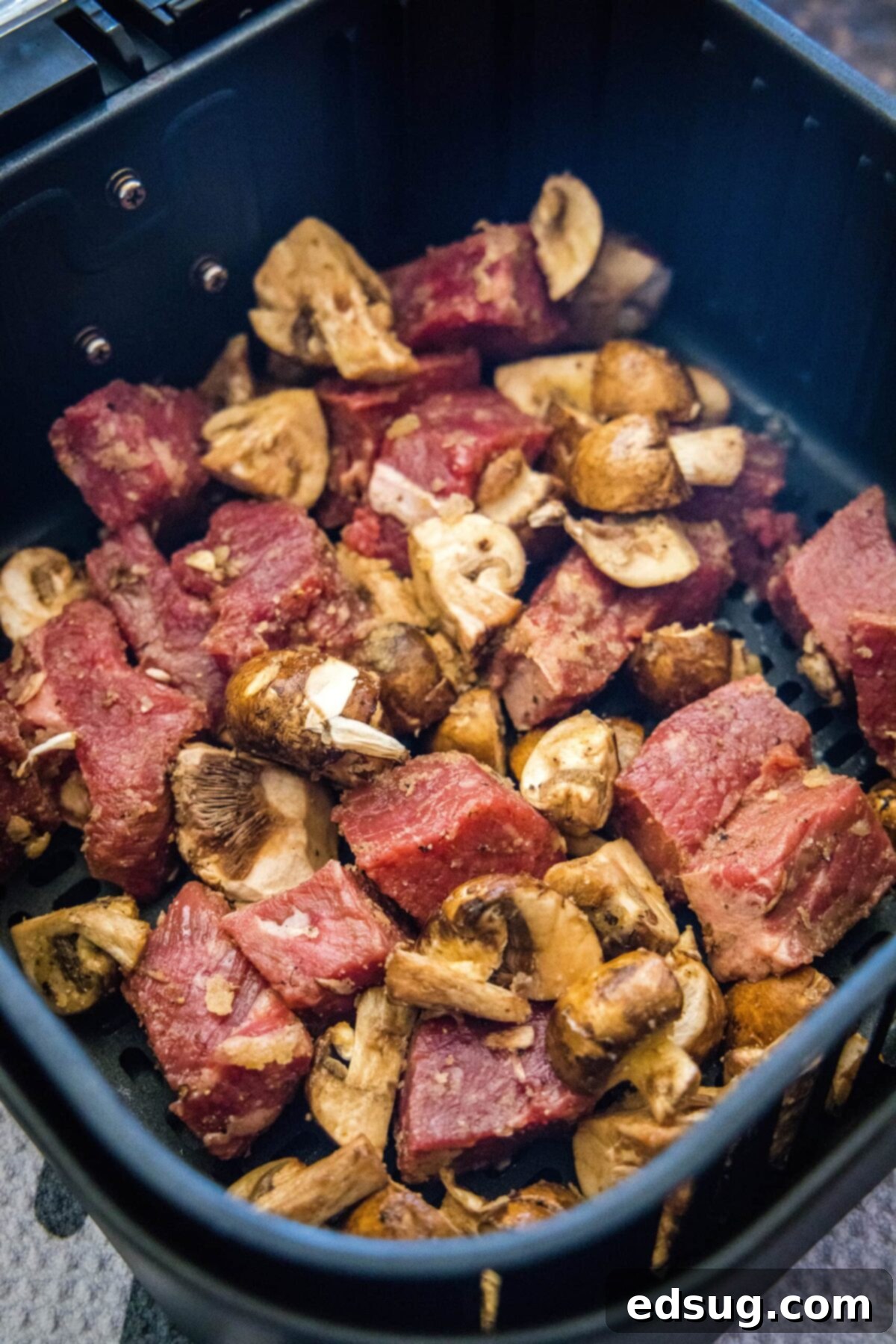 How to make easy air fryer steak bites with mushrooms and garlic powder, with tender meat and juicy mushrooms. It only takes 15 minutes! Perfectly seasoned, raw pieces of steak and quartered mushrooms arranged in a single, even layer within an air fryer basket, ready for cooking.