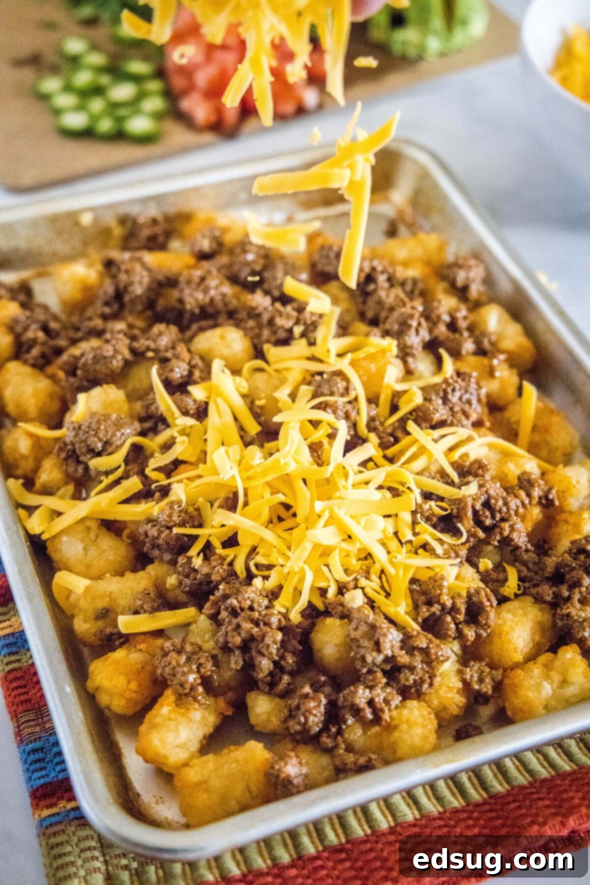 Shredded cheese being dropped onto a baking sheet with tater tots and ground beef