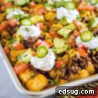The corner of a baking sheet with ground beef tater tot nachos, topped with jalapenos, sour cream, tomatoes, avocado, and cilantro
