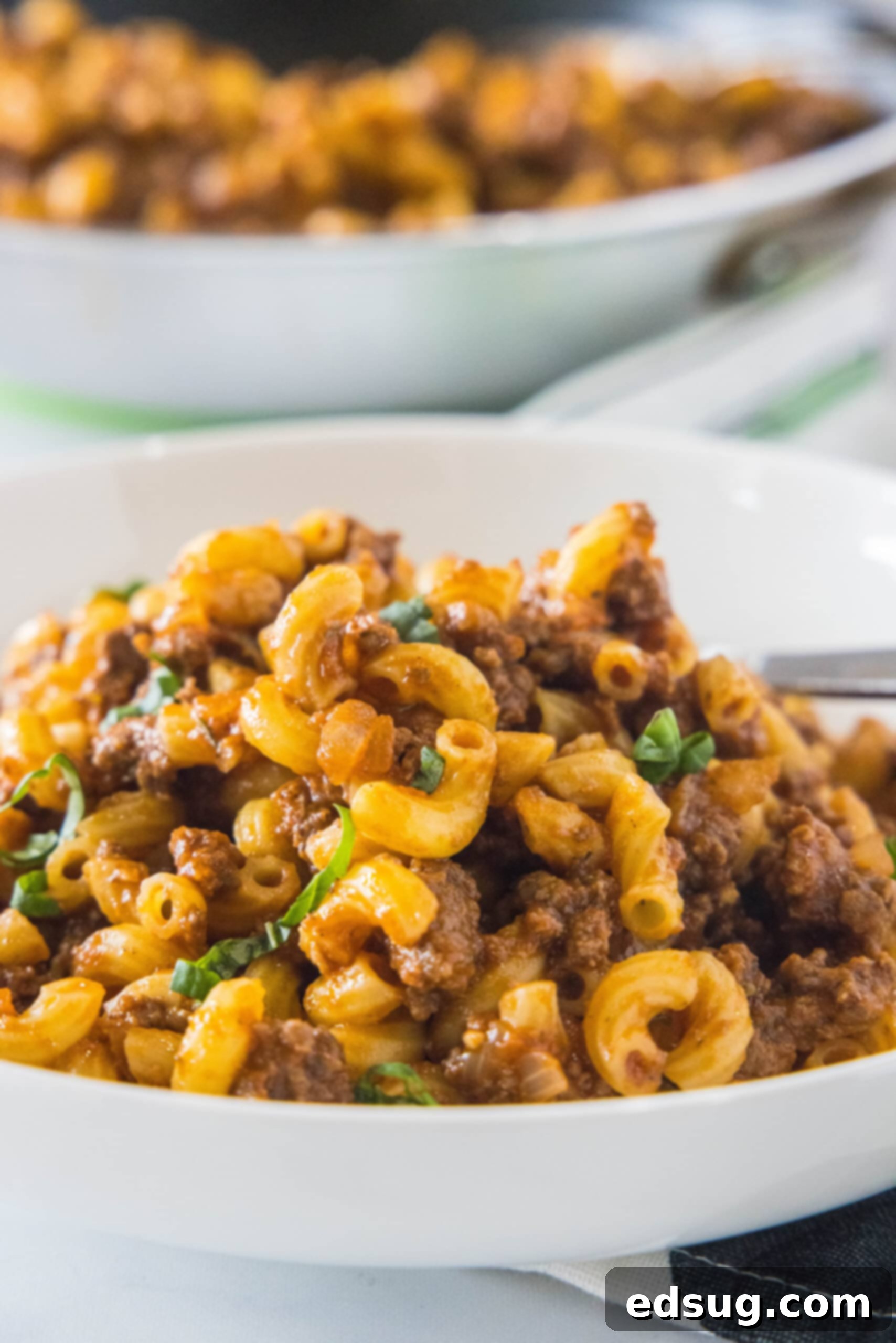 This easy and comforting homemade beefaroni recipe is ready in just 35 minutes, featuring savory ground beef, rich tomato sauce, and fragrant garlic. A steaming bowl of homemade beefaroni, with another blurred bowl in the background, showcasing its rich, comforting texture.