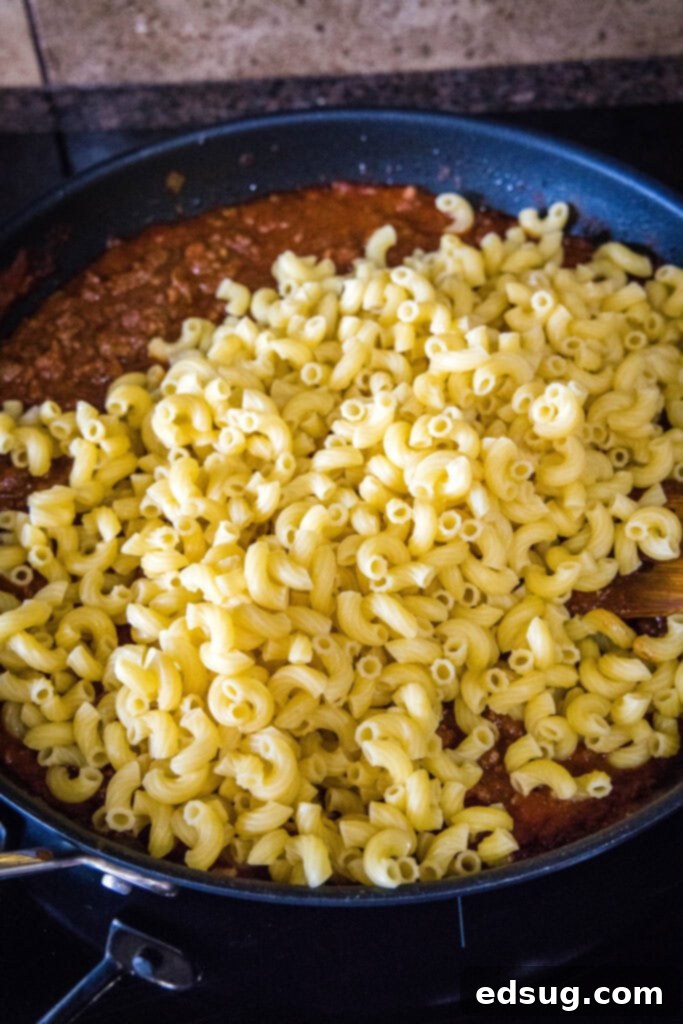 This step for easy beefaroni shows cooked elbow macaroni being added to the skillet of meat sauce, symbolizing the final combination of ingredients before serving. Freshly cooked and drained elbow macaroni being added to the rich, simmering beef and tomato sauce in a skillet, ready to be tossed and combined for the beefaroni.