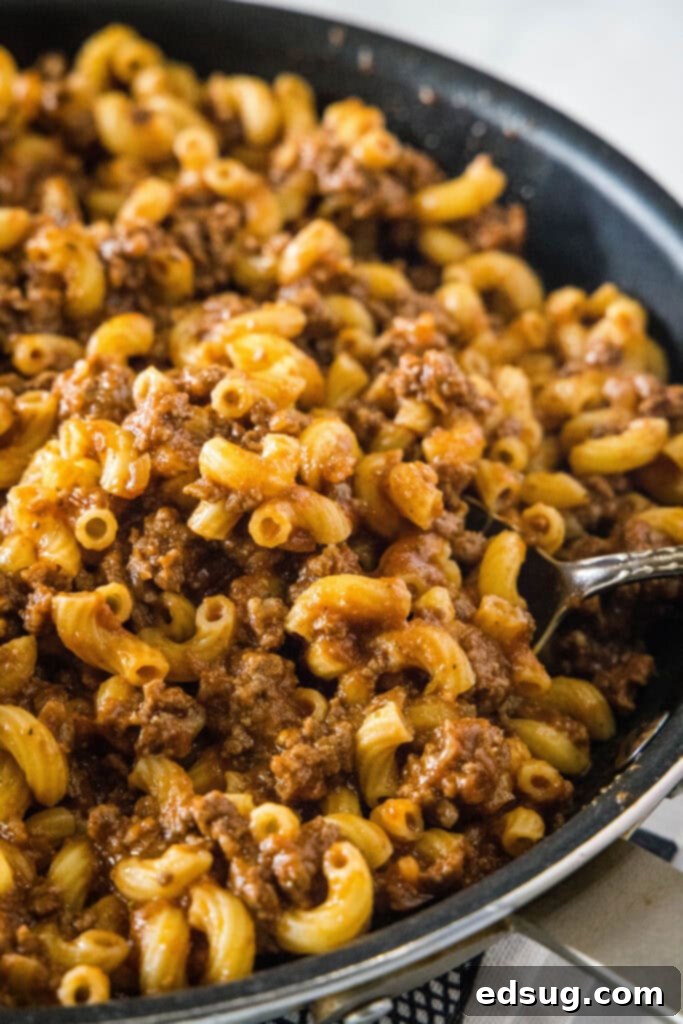 This image captures a delicious bite of beefaroni being lifted from the skillet, showcasing the completed dish ready to be enjoyed. A close-up shot of a fork lifting a bite of beefaroni from a skillet, showing the perfectly coated elbow macaroni mixed with the rich beef and tomato sauce, steam rising.