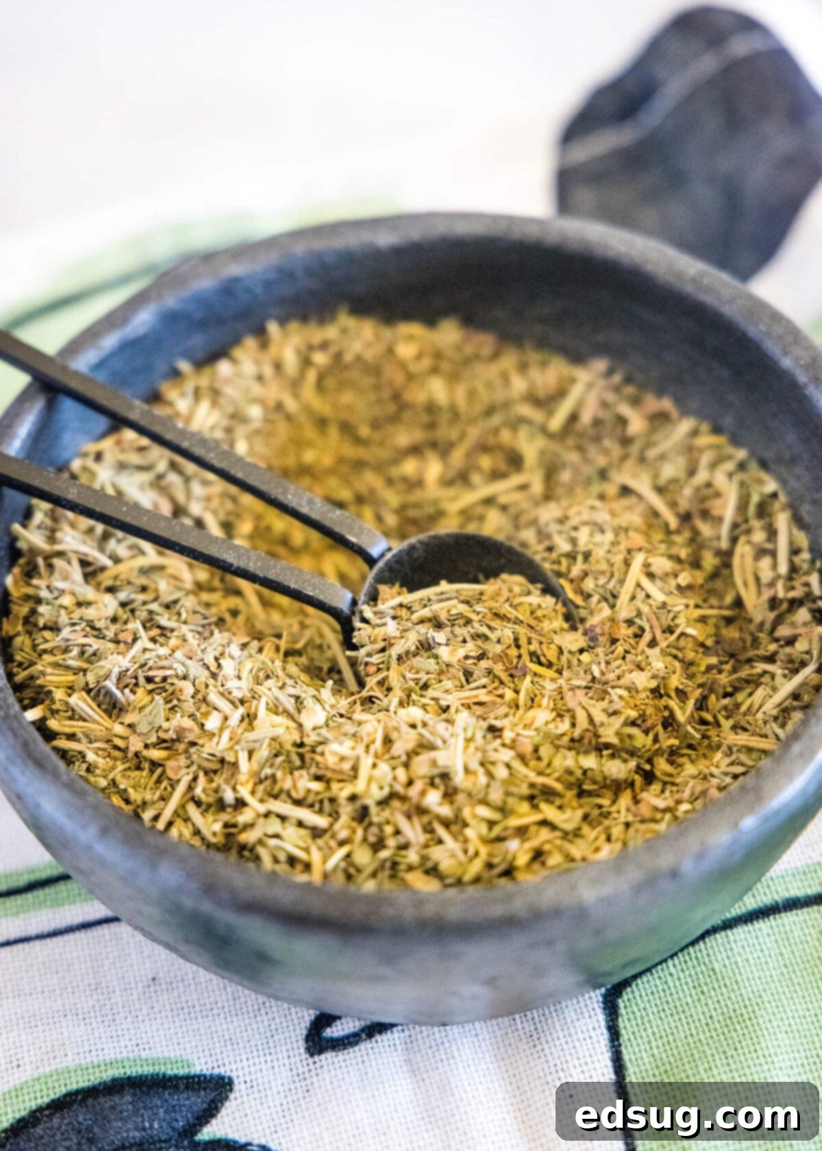 A close-up of a rustic bowl filled with vibrant homemade Italian seasoning, with a small wooden spoon resting inside.