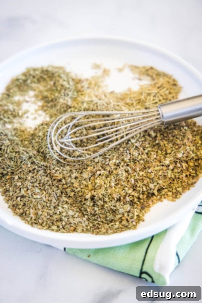 An overhead view captures a bowl of freshly mixed Italian seasoning, with a small whisk resting beside it on a white plate, highlighting the blend's texture and vibrant colors.