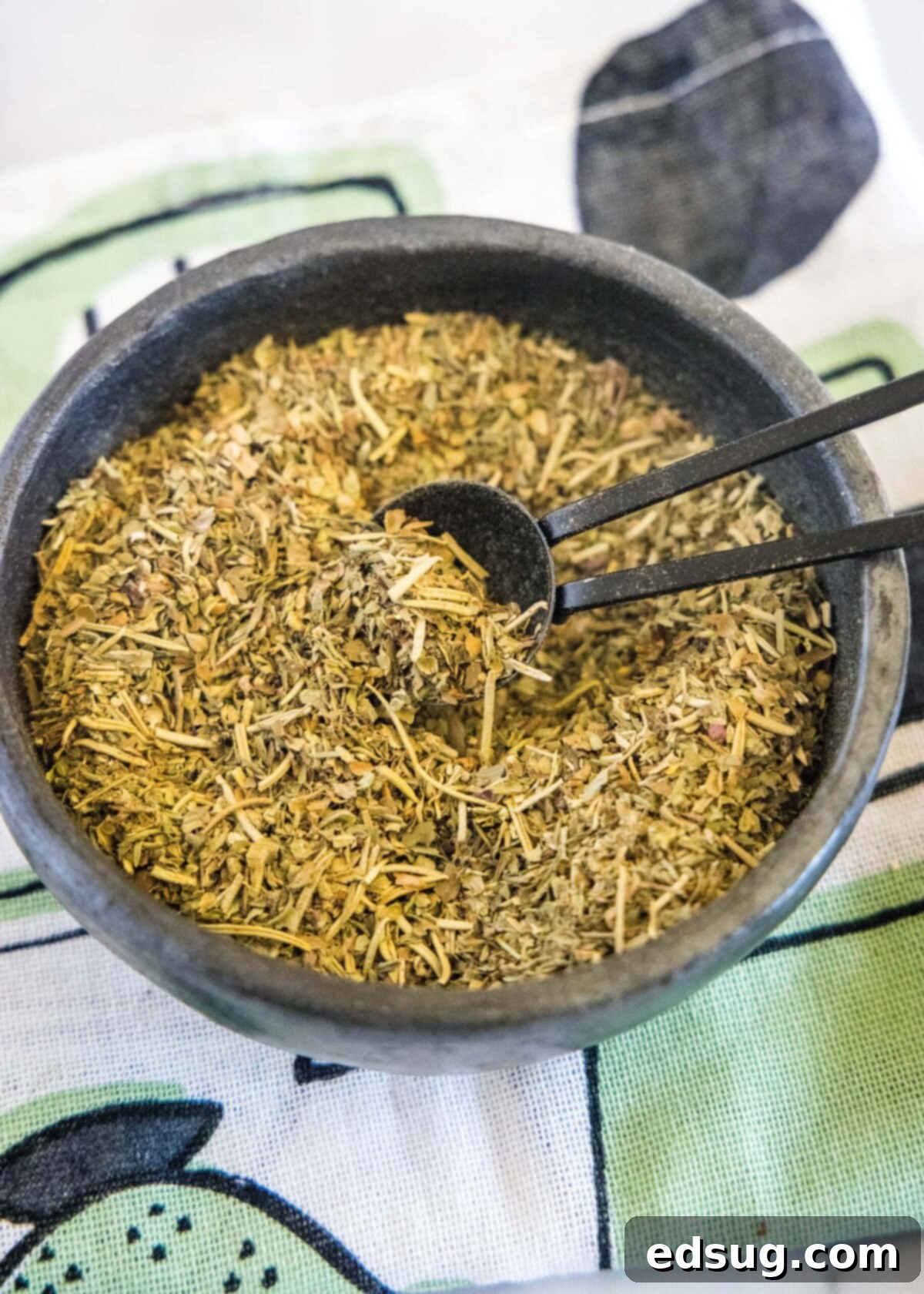 A close-up, overhead shot of a bowl of homemade Italian seasoning with a small wooden spoon, resting on a textured kitchen towel.