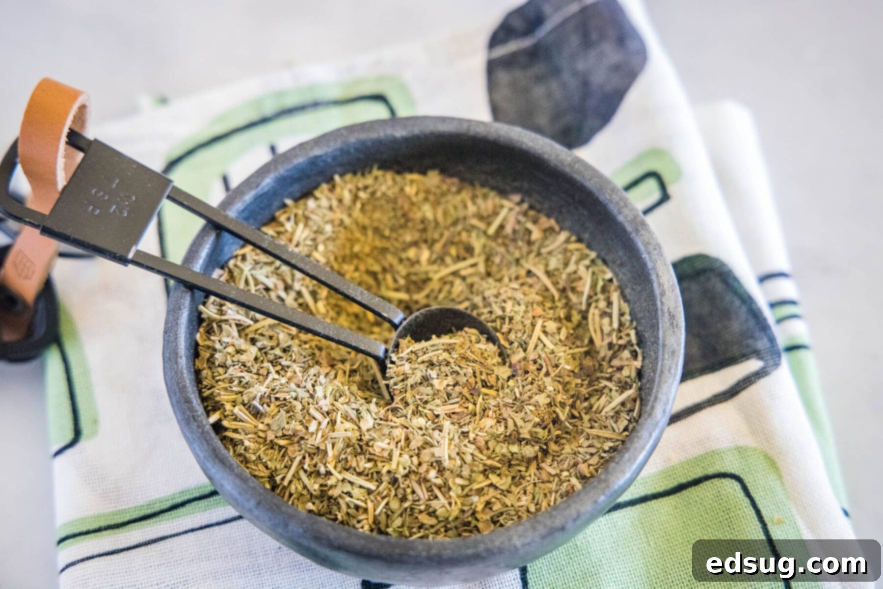 An inviting overhead view of a bowl filled with homemade Italian seasoning, accompanied by a wooden spoon, all set on a charming kitchen towel.
