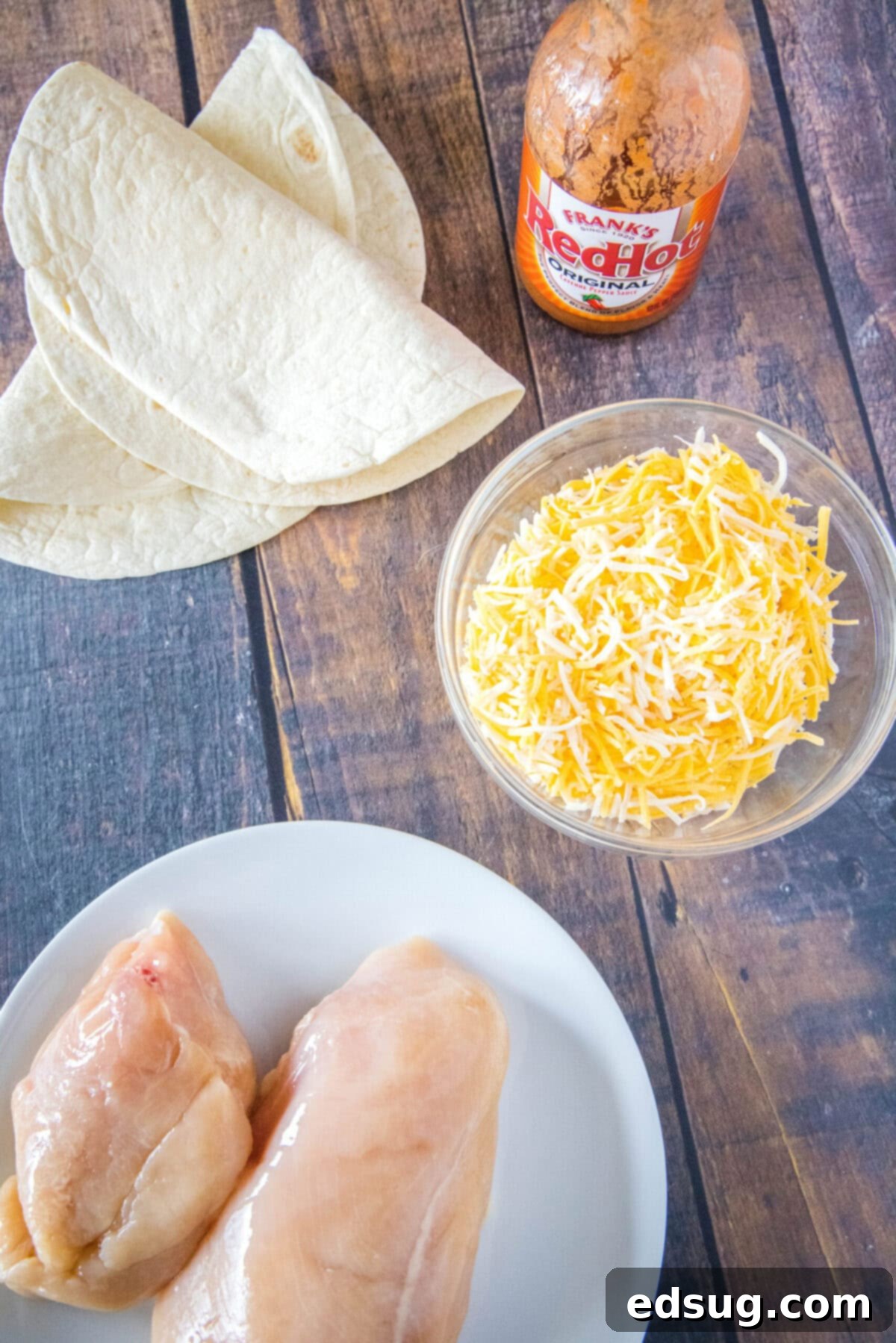 This easy homemade buffalo chicken quesadilla recipe is loaded with juicy chicken, melty cheese, and spicy wing sauce. Overhead view of a plate of chicken breasts, a bowl of shredded cheese, a bottle of wing sauce, and two flour tortillas