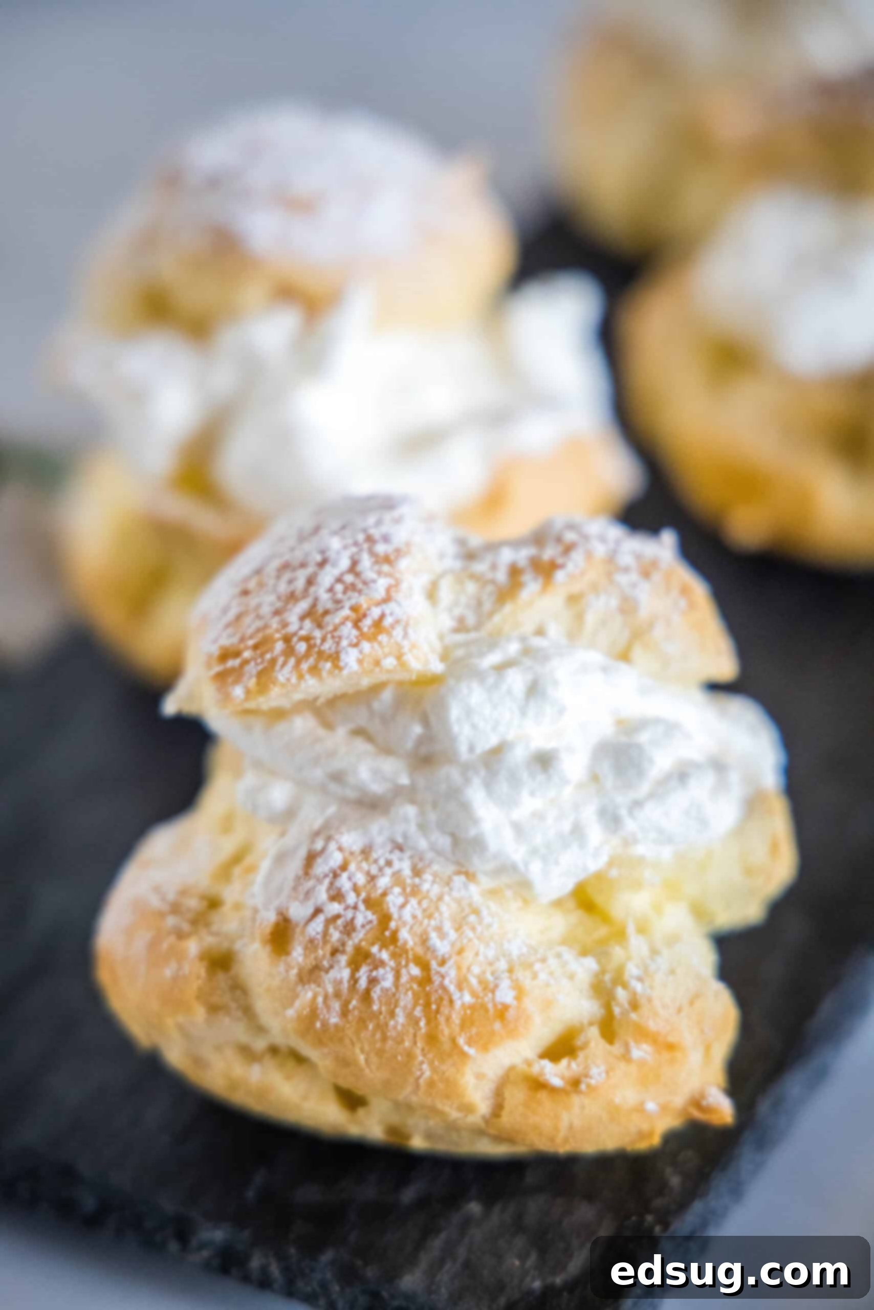 Learn how to make perfectly light and flaky homemade cream puffs (profiteroles) from scratch using an easy choux pastry recipe, filled with sweet whipped cream and dusted with powdered sugar. Close up of a homemade cream puff dusted with powdered sugar, with more cream puffs blurred in the background on a platter.