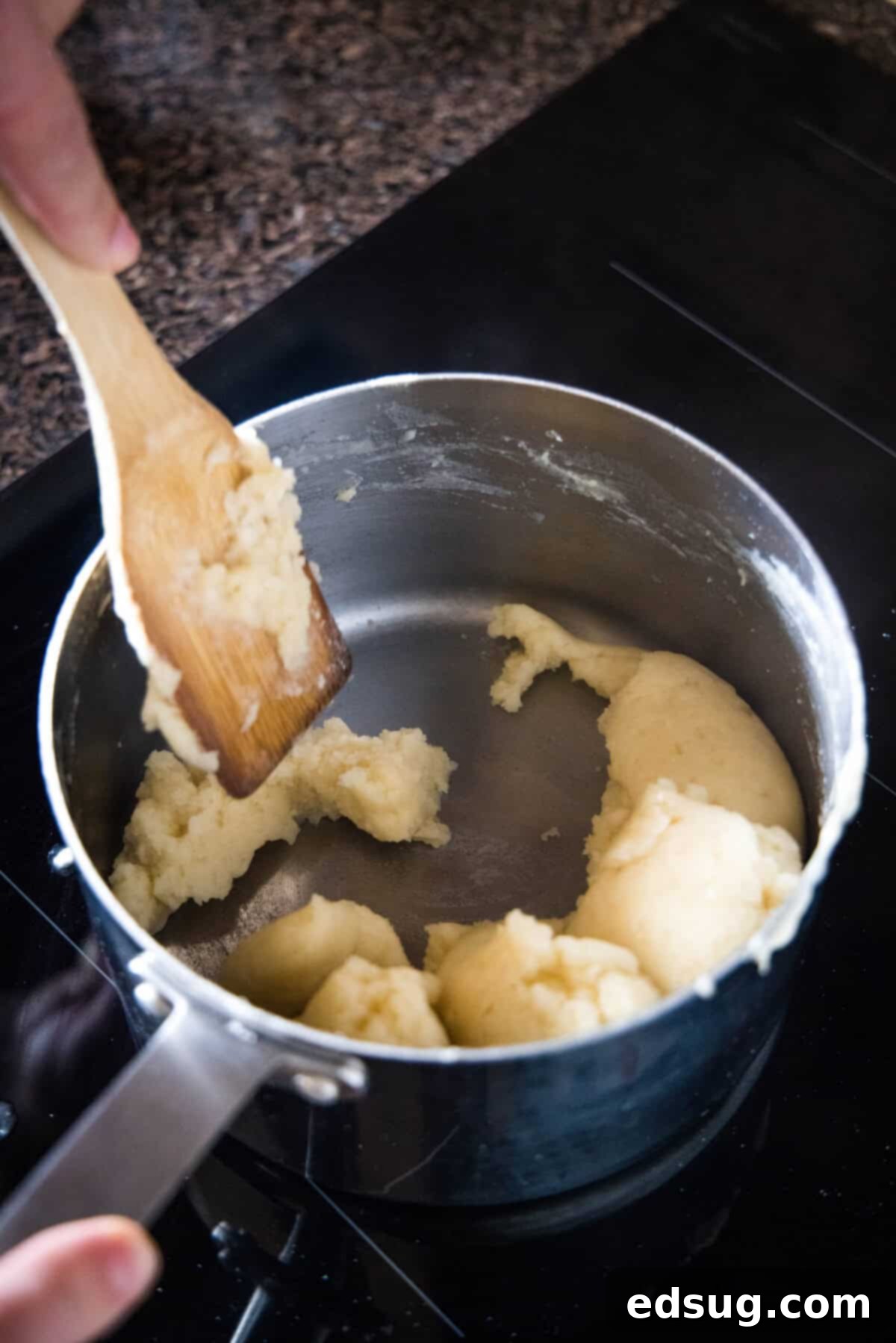 Stir the choux pastry dough until it forms a ball and pulls away from the pan. A wooden spoon stirring butter and flour mixture vigorously inside a saucepan to form a smooth dough.