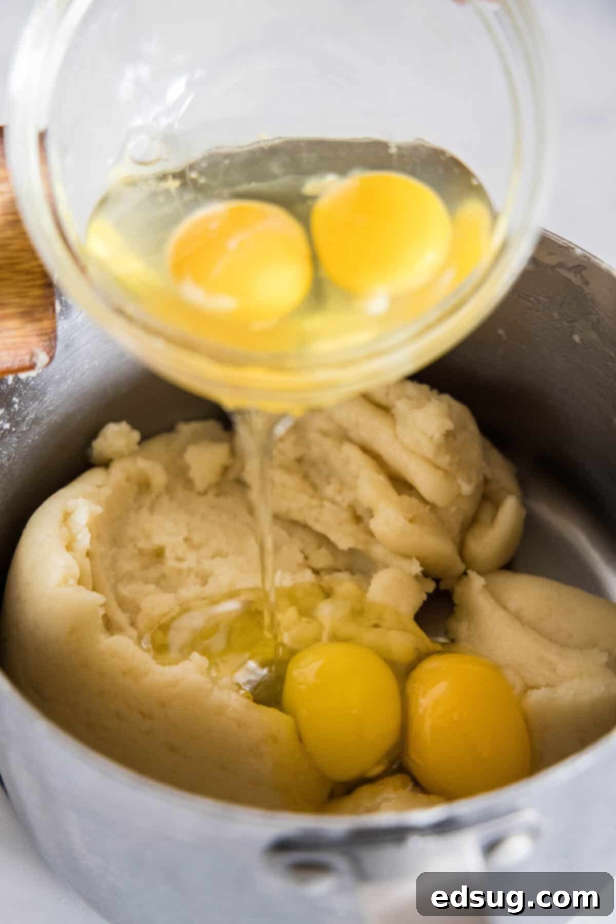 Beat eggs into the warm choux pastry dough until smooth. Cracked eggs being added one by one into the warm choux pastry dough in a saucepan.