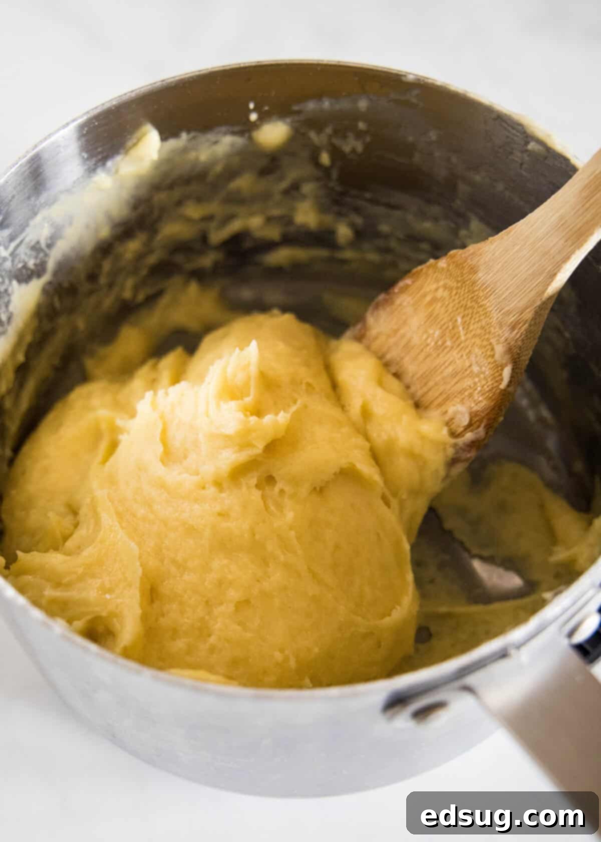 Continue beating until the choux pastry forms a thick, smooth batter. A wooden spoon mixing the choux pastry dough in a saucepan after eggs have been added, resulting in a thick, smooth batter.