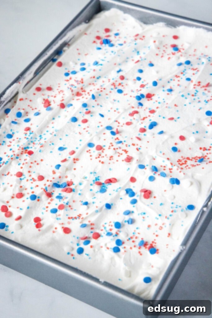 Frosted patriotic poke cake topped with fluffy white whipped cream and festive red, white, and blue sprinkles