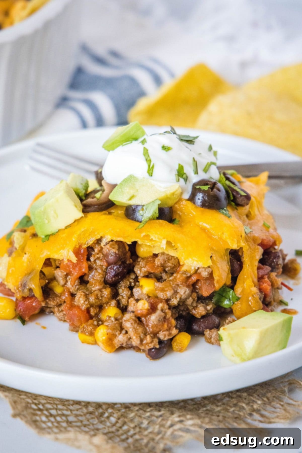 This easy 30-minute taco casserole is loaded with juicy and tender beef, gooey melted cheese, and your favorite taco toppings. A plate with a serving of hearty taco casserole, beautifully topped with fresh sour cream, bright cilantro, black olives, and creamy avocado slices, with a bowl of crunchy tortilla chips in the background.