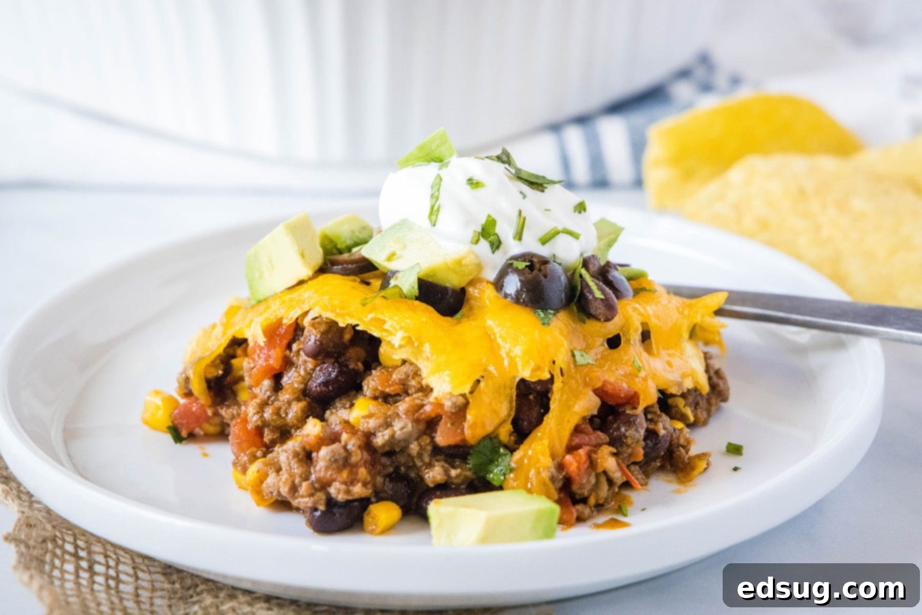 This easy 30-minute taco casserole is loaded with juicy and tender beef, gooey melted cheese, and your favorite taco toppings. A close-up of a serving of taco casserole on a white plate, ready to eat, garnished with sour cream, cilantro, black olives, and avocado slices. A fork is resting beside it.
