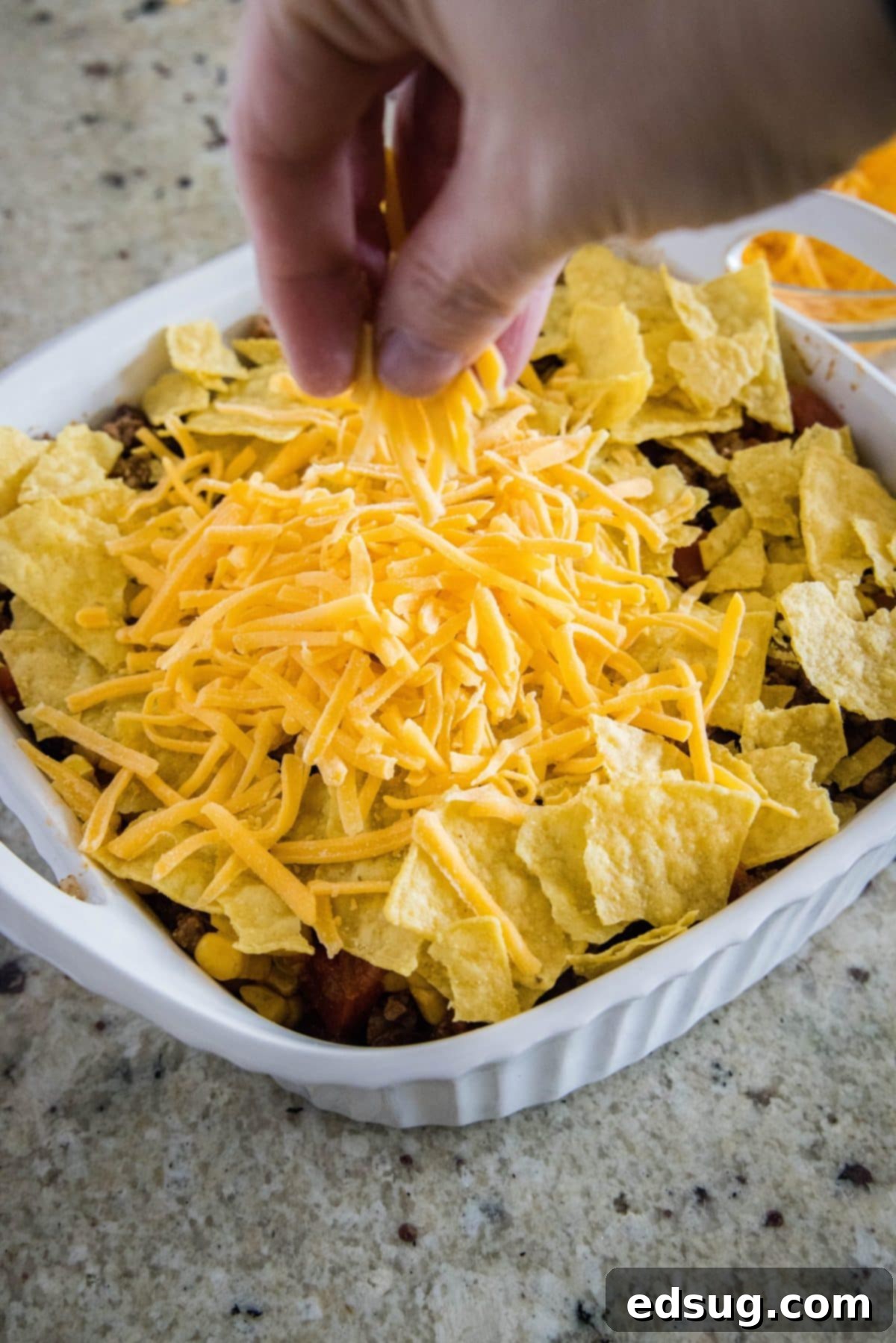This easy 30-minute taco casserole is loaded with juicy and tender beef, gooey melted cheese, and your favorite taco toppings. A hand gently sprinkling a generous amount of shredded cheddar cheese over a layer of crunchy tortilla chips in a baking dish, preparing the casserole for baking.
